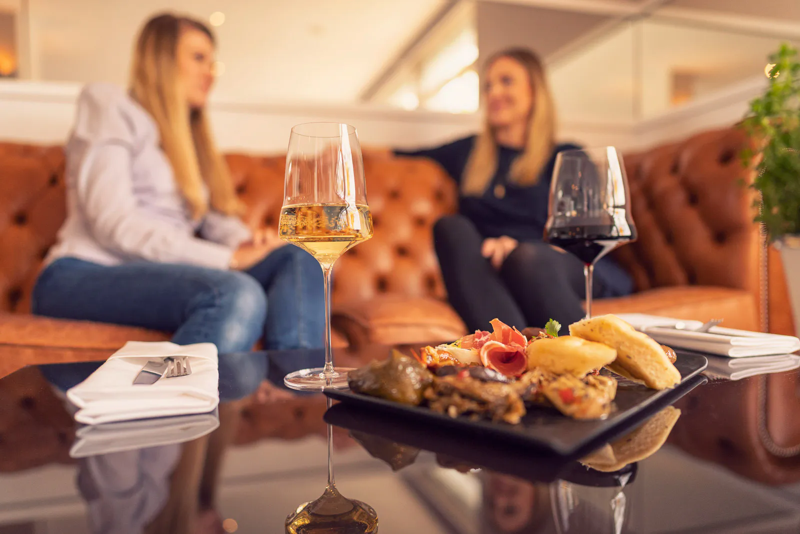Zwei Frauen sitzen auf einem braunen Ledersofa, davor stehen Weingläser und eine Platte mit Speisen im warmen Licht.