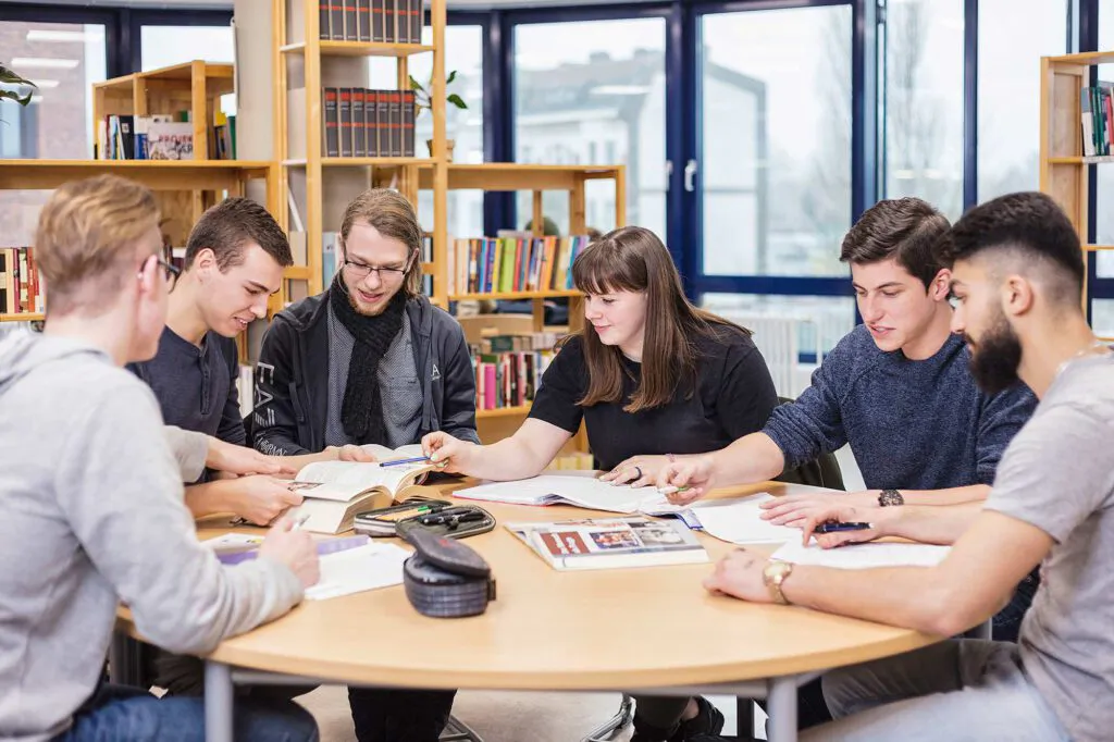 Sechs junge Erwachsene lernen gemeinsam an einem Tisch mit Büchern in einer hellen Bibliothek.