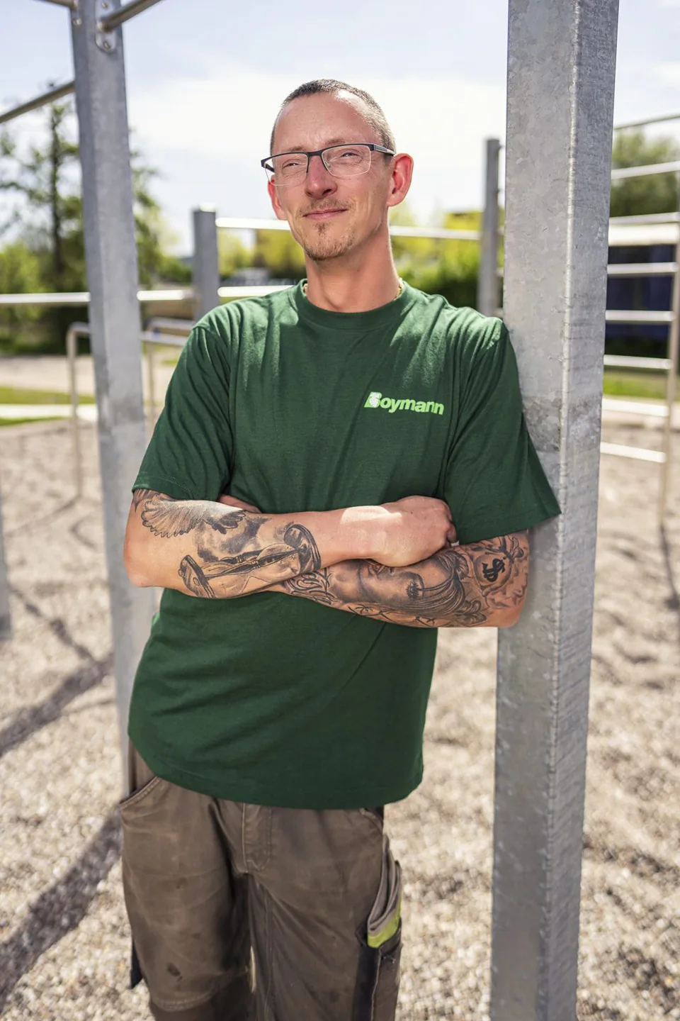 Mann mit Brille und tätowierten Armen im grünen Boymann-Shirt lehnt bei Sonnenlicht an einem Metallpfosten im Freien.