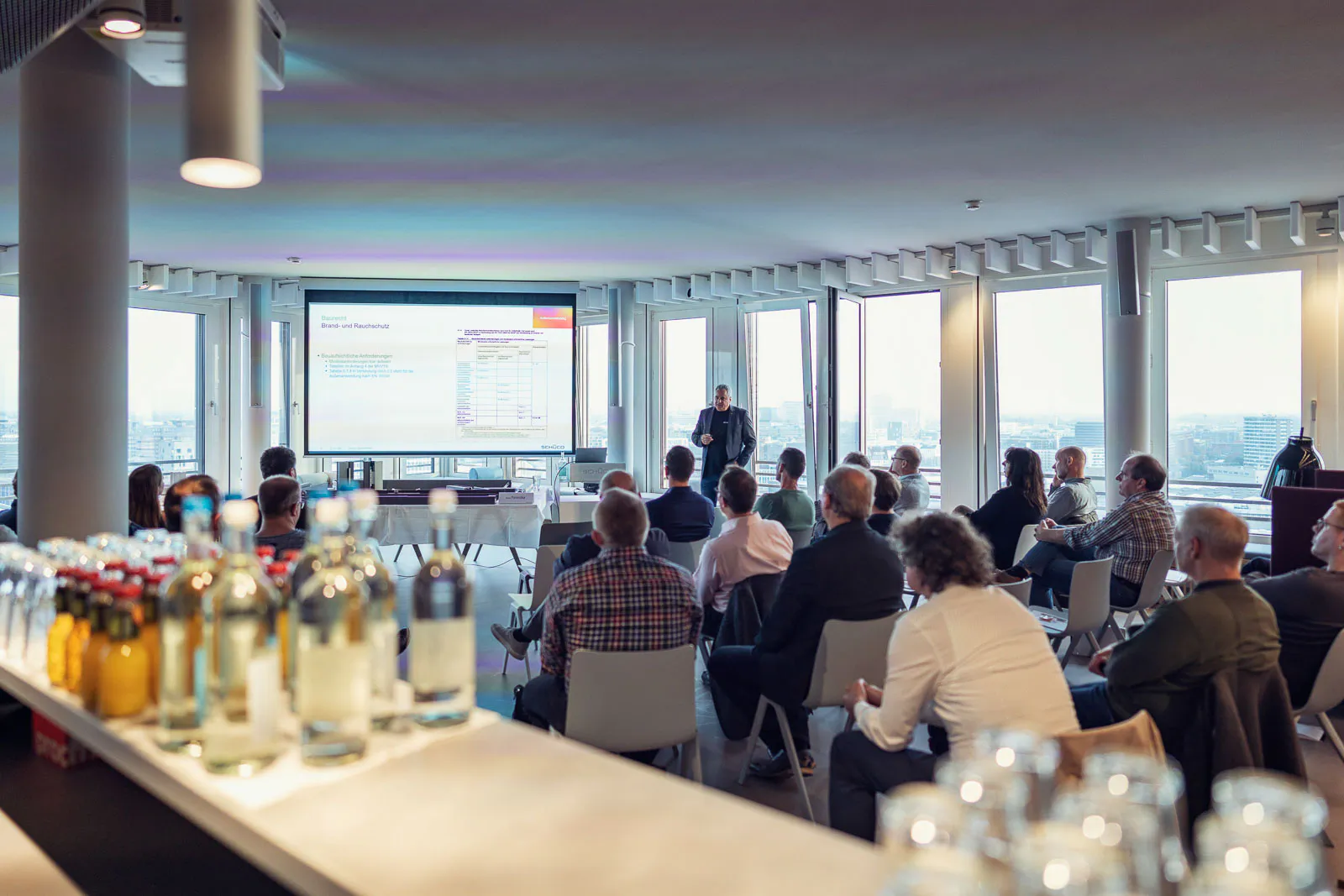 Publikum hört einem Vortragenden neben einer Leinwand im hellen Veranstaltungsraum zu, Getränkeflaschen im Vordergrund.