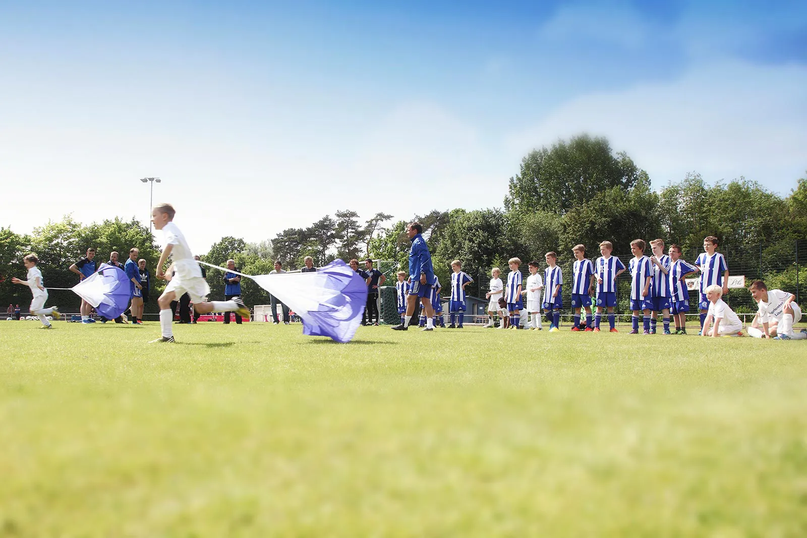 Kinder tragen Fahnen über den sonnigen Fußballplatz, dahinter stehen Spieler in blau-weißen Trikots.