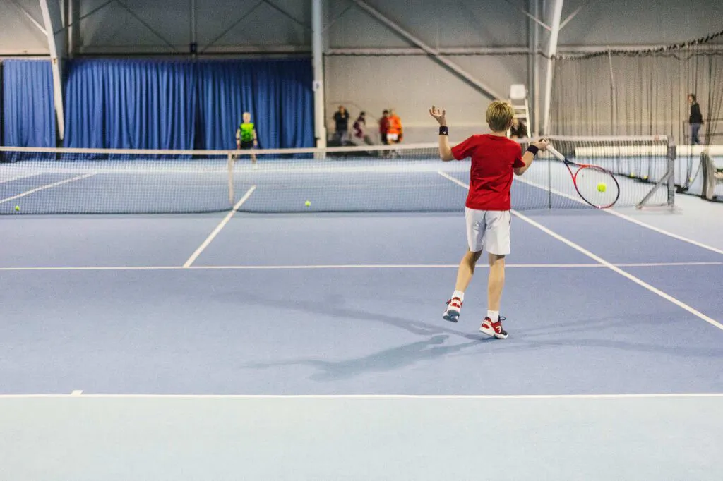 Junger Tennisspieler in rotem Shirt schlägt beim Wilson Master Cup einen Ball auf blauem Hallenplatz.