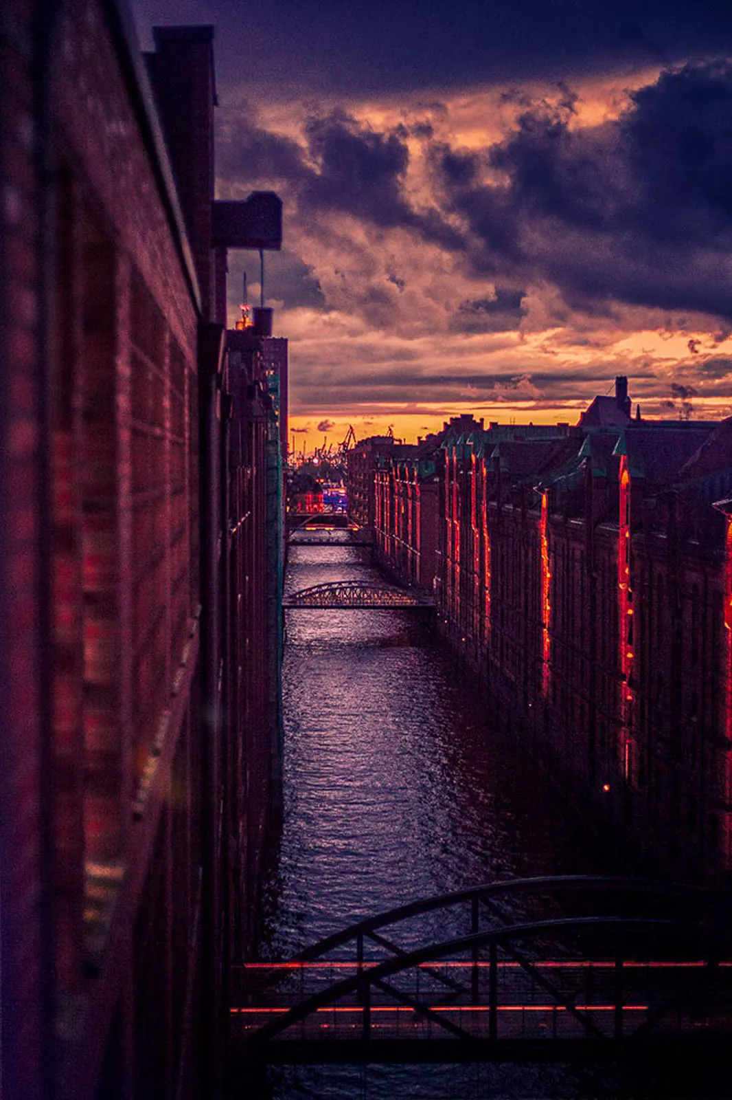 Schmaler Kanal zwischen Backsteinspeichern mit Brücken im rötlichen Abendlicht unter dunklen Wolken.