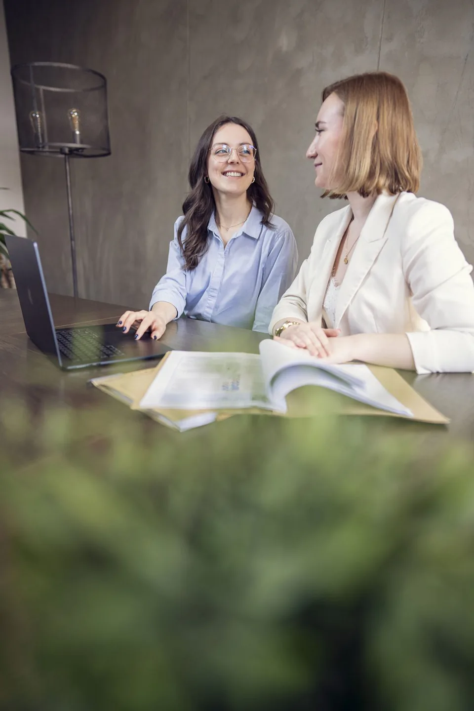 Zwei Frauen sitzen im Büro am Tisch mit Laptop und aufgeschlagenem Katalog und lächeln im Gespräch.
