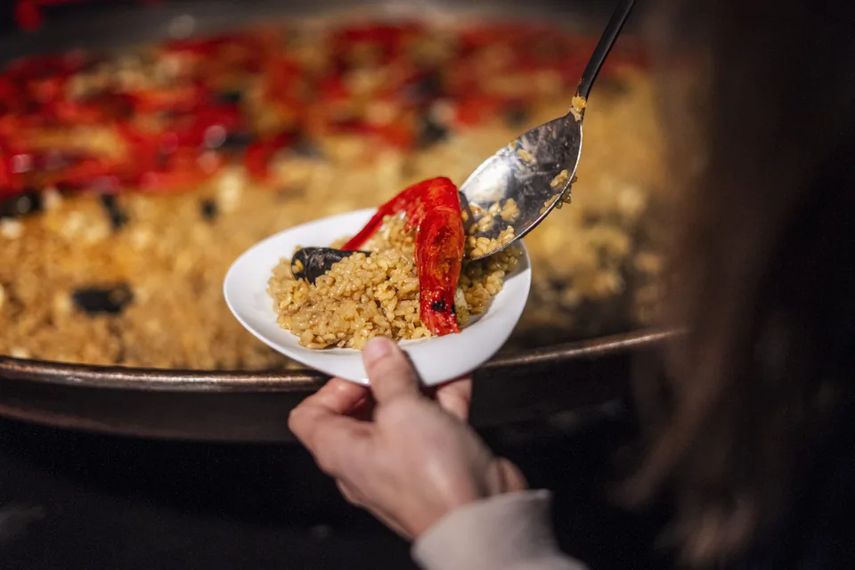 Eine Person schöpft Reis mit roter Paprika und Muscheln aus einer großen Pfanne auf einen weißen Teller.