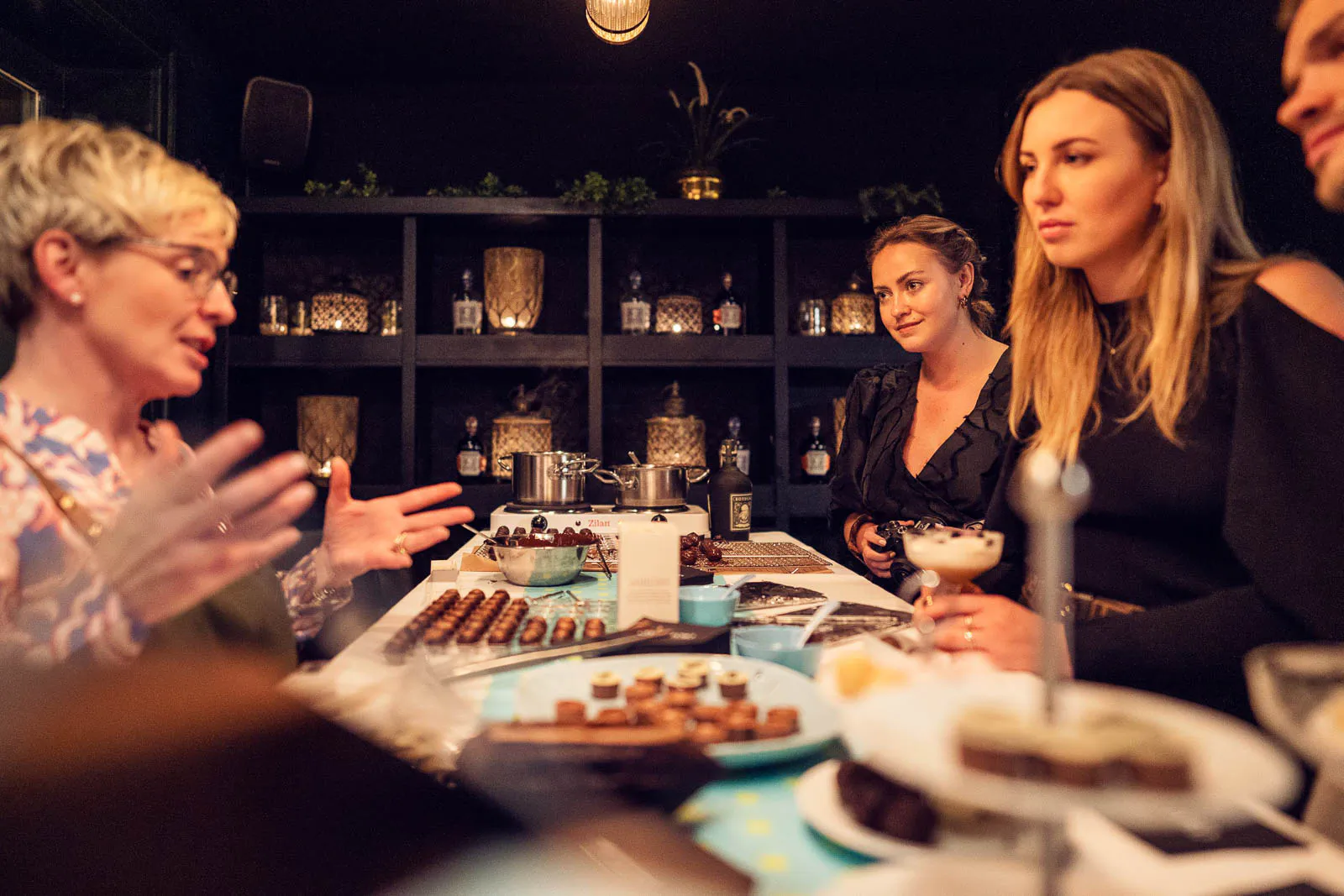 Gäste an einer Bar mit Pralinen, Dessertgläsern und Flaschenregalen bei warmem Licht beim BOTUCAL-Event.