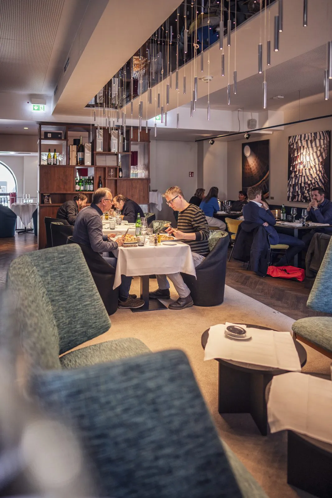 Gäste speisen an weiß gedeckten Tischen in einem Restaurant mit Polstersesseln und filigranen Pendelleuchten.