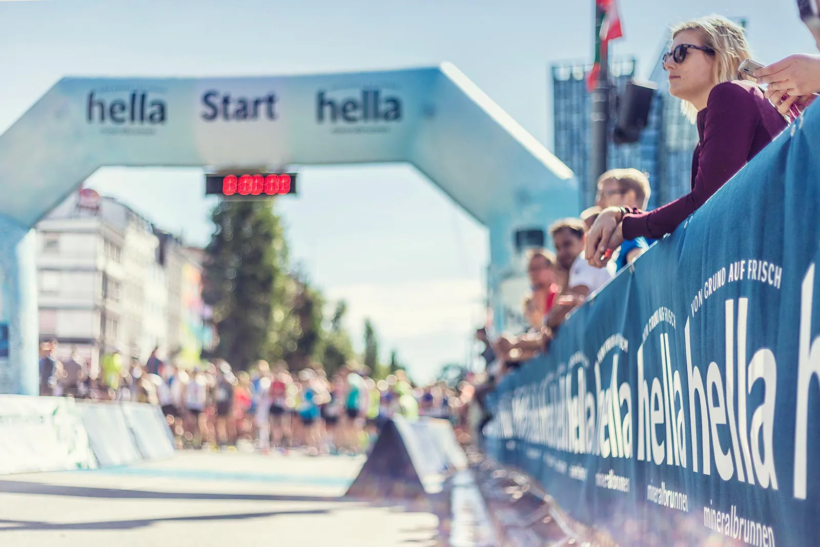 Zuschauer lehnen an blauen Absperrungen neben dem Startbogen des Hella Halbmarathons im Sonnenlicht.