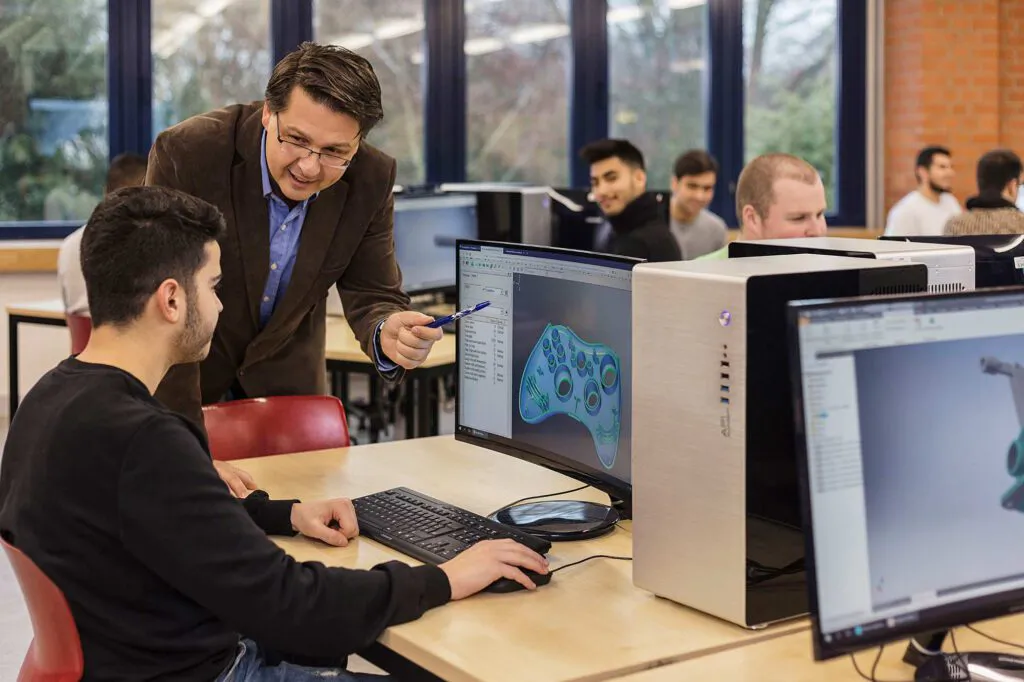 Ein Lehrer zeigt einem Schüler im hellen Computerraum ein 3D-Modell eines Controllers auf dem Monitor.