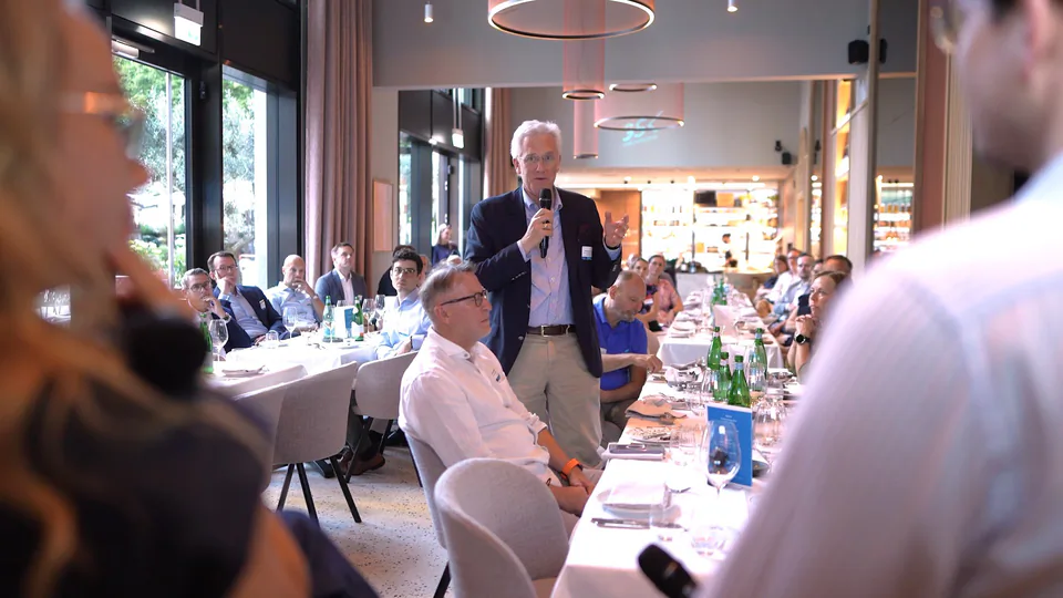 Ein Mann spricht mit Mikrofon zwischen gedeckten Tischen vor sitzenden Gästen in einem hellen Veranstaltungsraum.