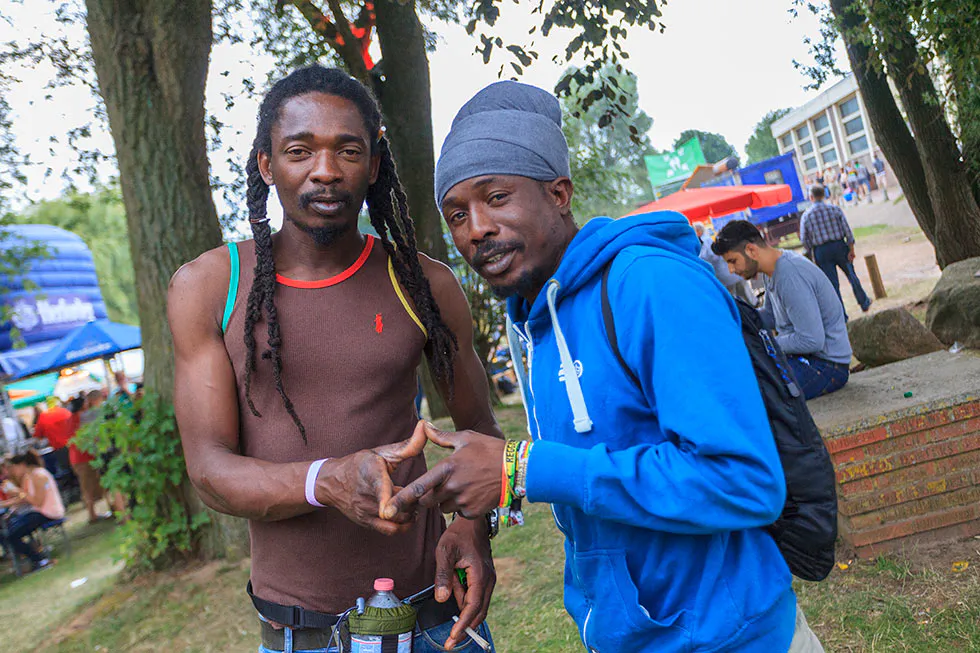 Zwei Männer posieren unter Bäumen auf dem Festivalgelände, einer mit langen Zöpfen, der andere im blauen Hoodie.