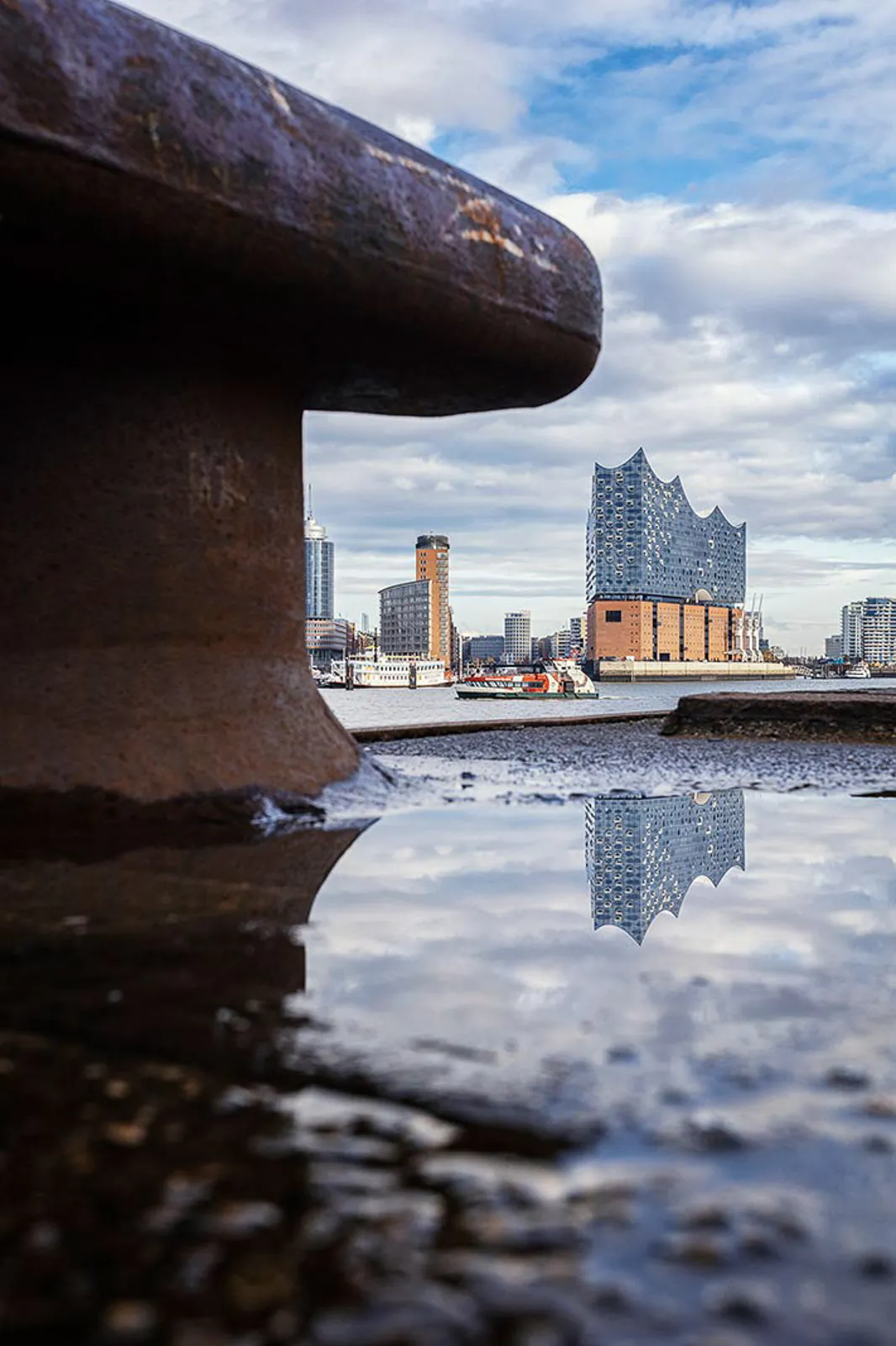 Elbphilharmonie am Wasser unter bewölktem Himmel, in einer Pfütze gespiegelt; großer rostiger Poller im Vordergrund.