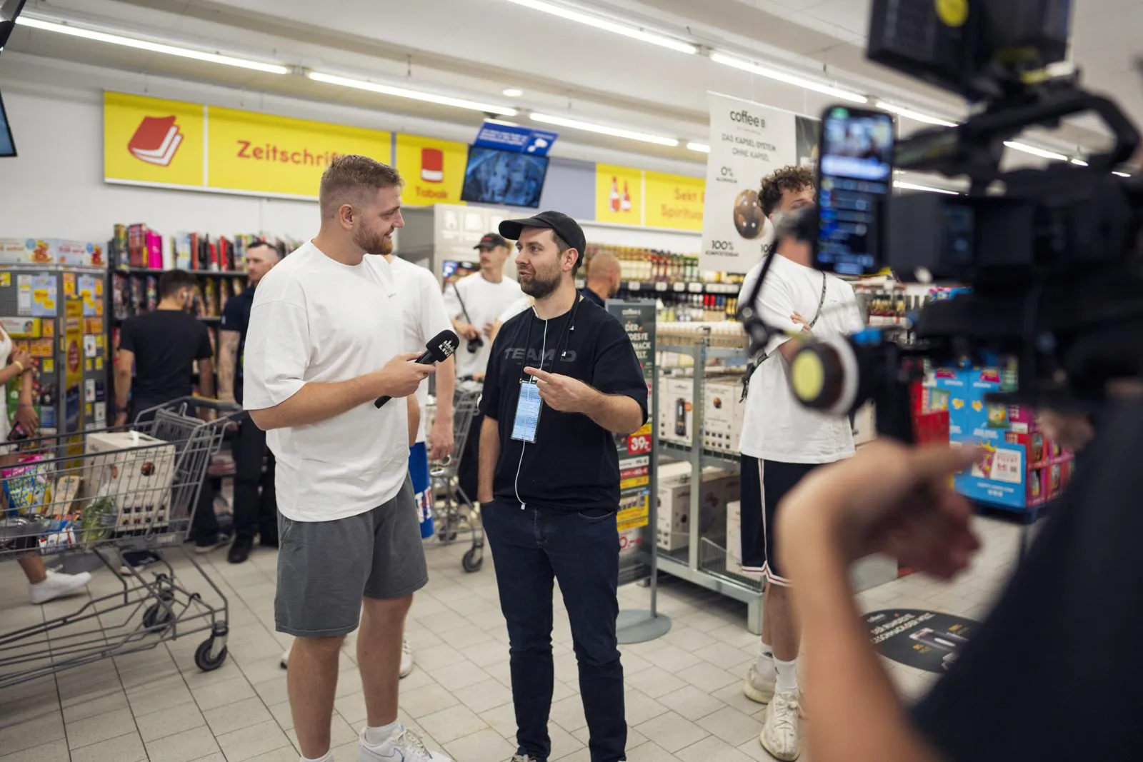 Zwei Männer beim Interview im hell beleuchteten Netto-Markt, eine Videokamera im Vordergrund.