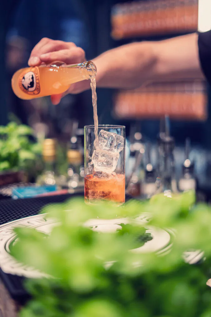 Eine Hand gießt ein orangefarbenes Getränk aus einer Glasflasche in ein hohes Glas mit Eis; Kräuter stehen im Vordergrund.