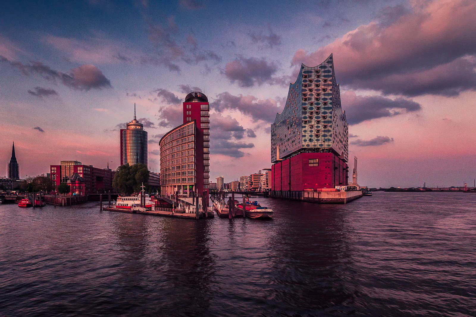 Elbphilharmonie am Wasser unter rosa Abendwolken, daneben Backsteingebäude und Boote am Anleger.