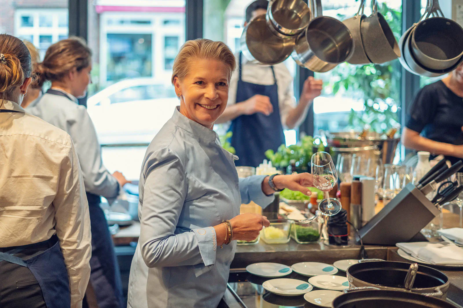 Cornelia Poletto lächelt mit einem Weinglas in der Hand in einer hellen Restaurantküche mit hängenden Töpfen.