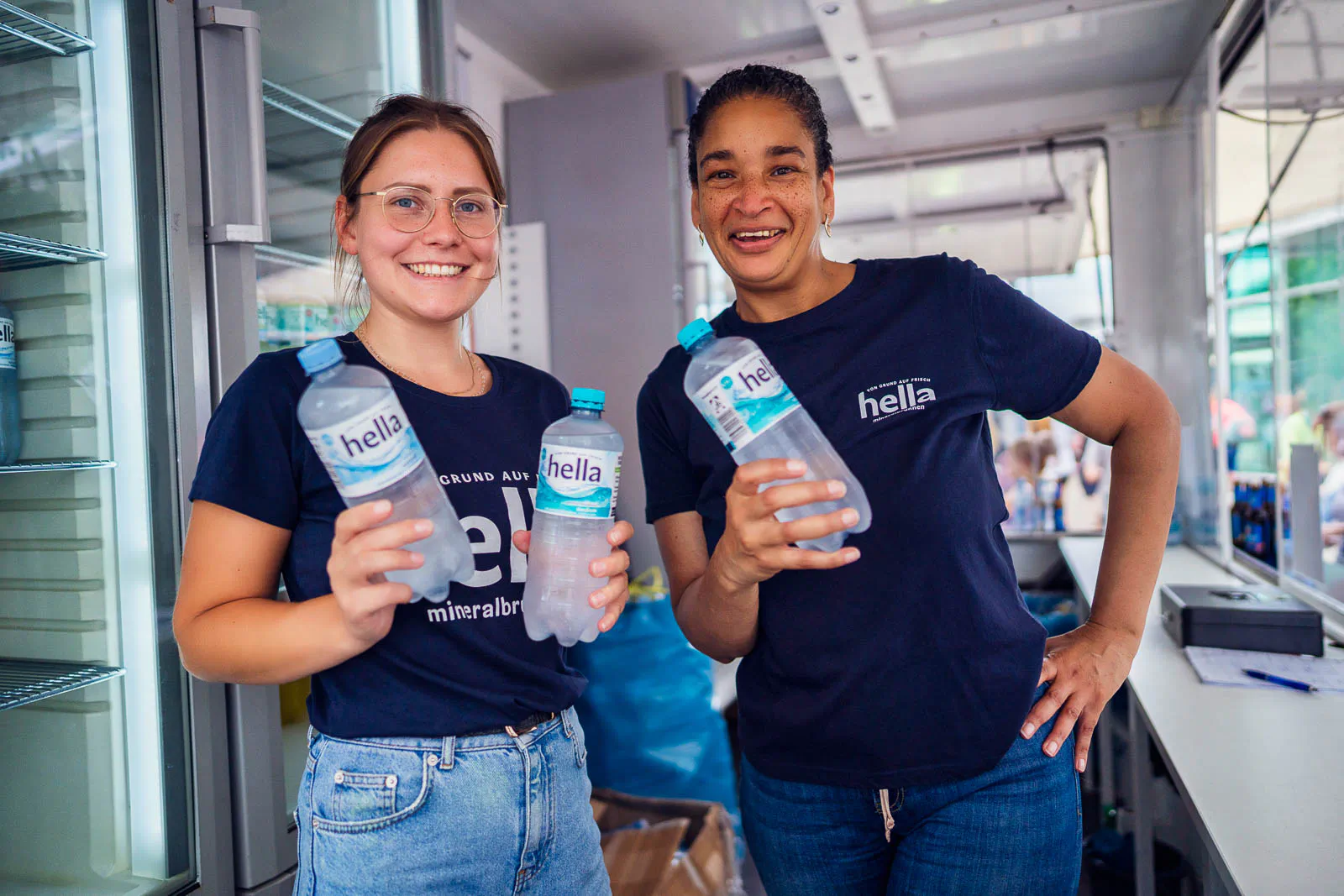 Zwei lächelnde Frauen in dunkelblauen Hella-Shirts halten Wasserflaschen in einem hellen Getränkestand.