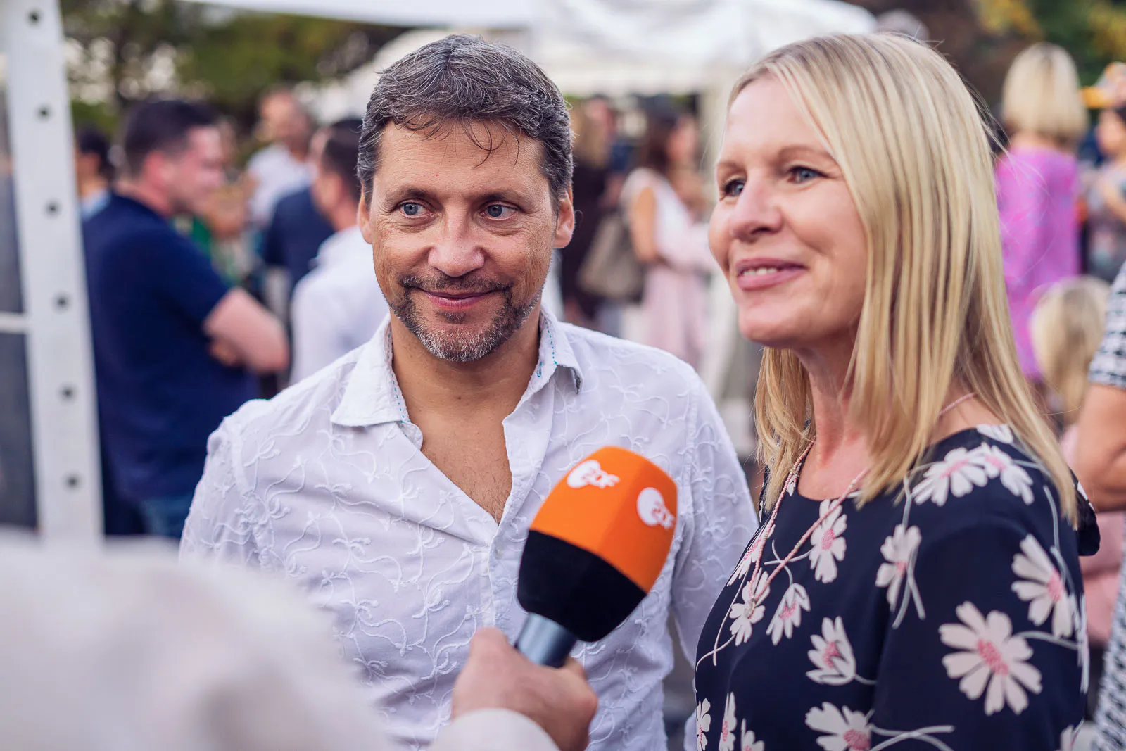 Mann und blonde Frau beim Interview mit orangefarbenem Mikrofon auf einer Sommerparty im Freien bei Tageslicht.