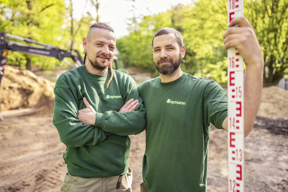 Zwei Männer in grünen Boymann-Shirts auf einer sonnigen Baustelle, einer hält eine Messlatte, dahinter ein Bagger.