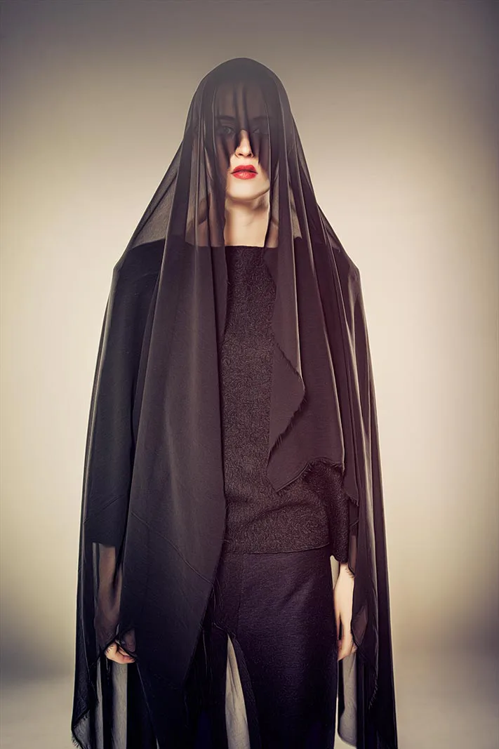 Person in schwarzem Outfit mit transparentem Schleier über dem Kopf und roten Lippen vor warm beleuchtetem Studiohintergrund.
