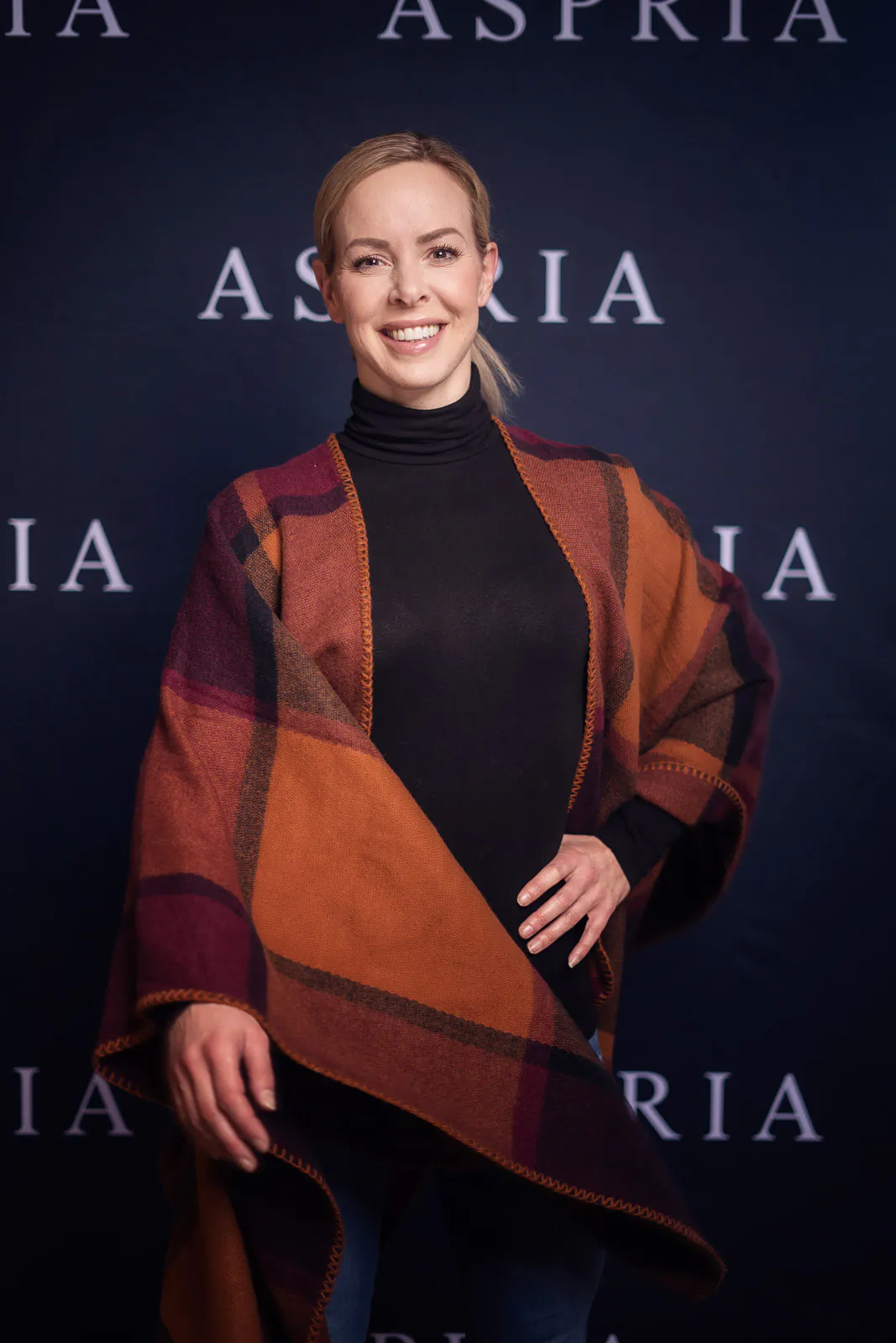 Isabel Edvardsson lächelt im schwarzen Rollkragenpullover und orange karierten Poncho vor einer Aspria-Logowand.