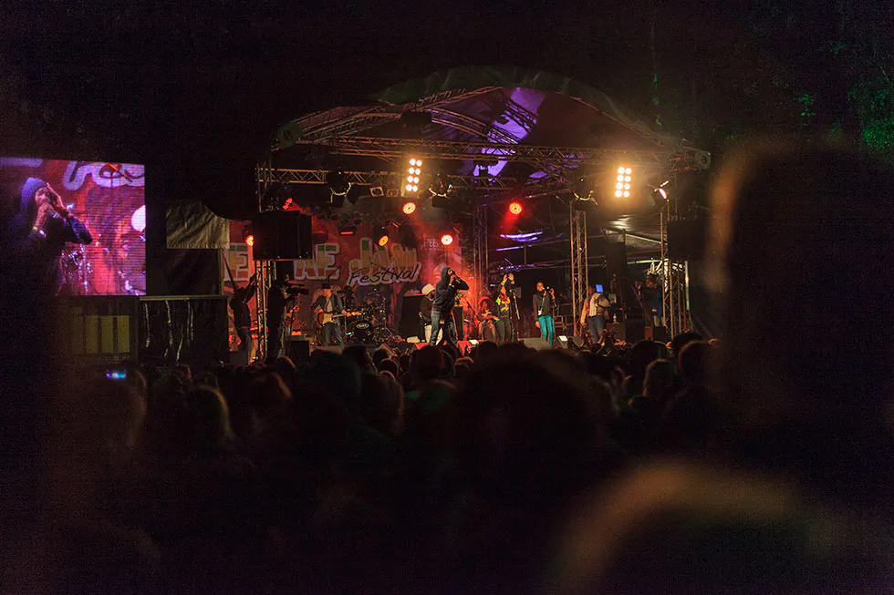 Publikum vor einer überdachten Konzertbühne mit Band, roten und gelben Scheinwerfern und Videoleinwand bei Nacht.