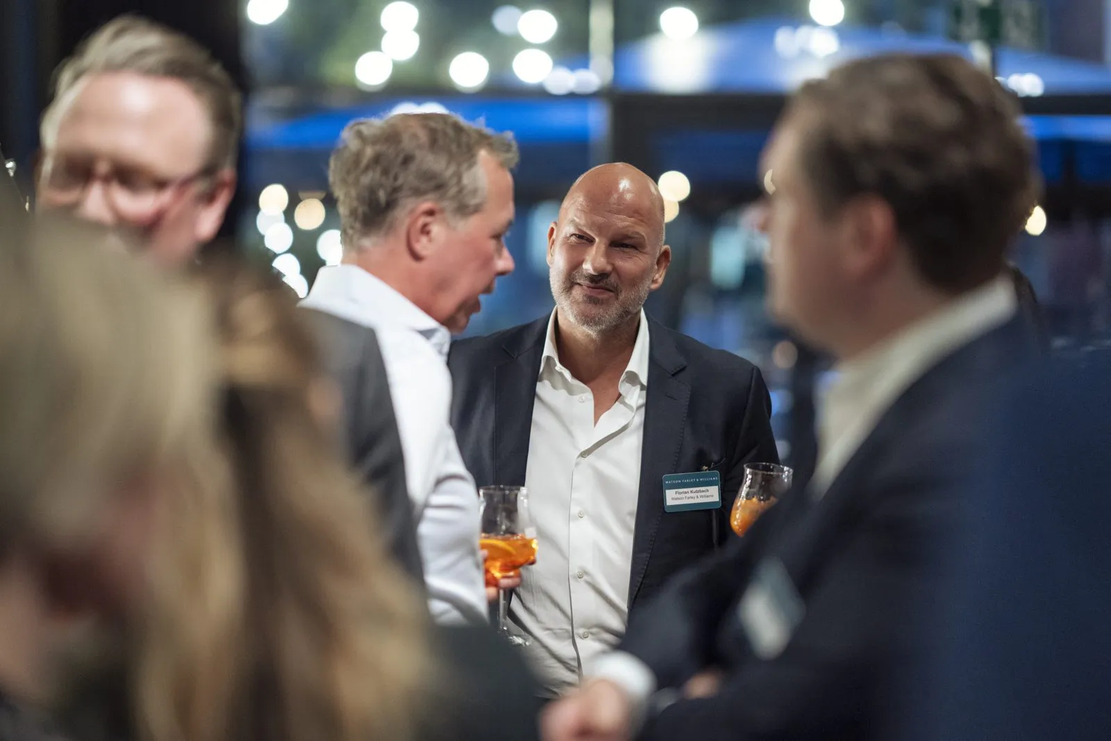 Gäste in Anzügen unterhalten sich mit Getränken bei einem Watson & Farley-Event im warm beleuchteten Innenraum.