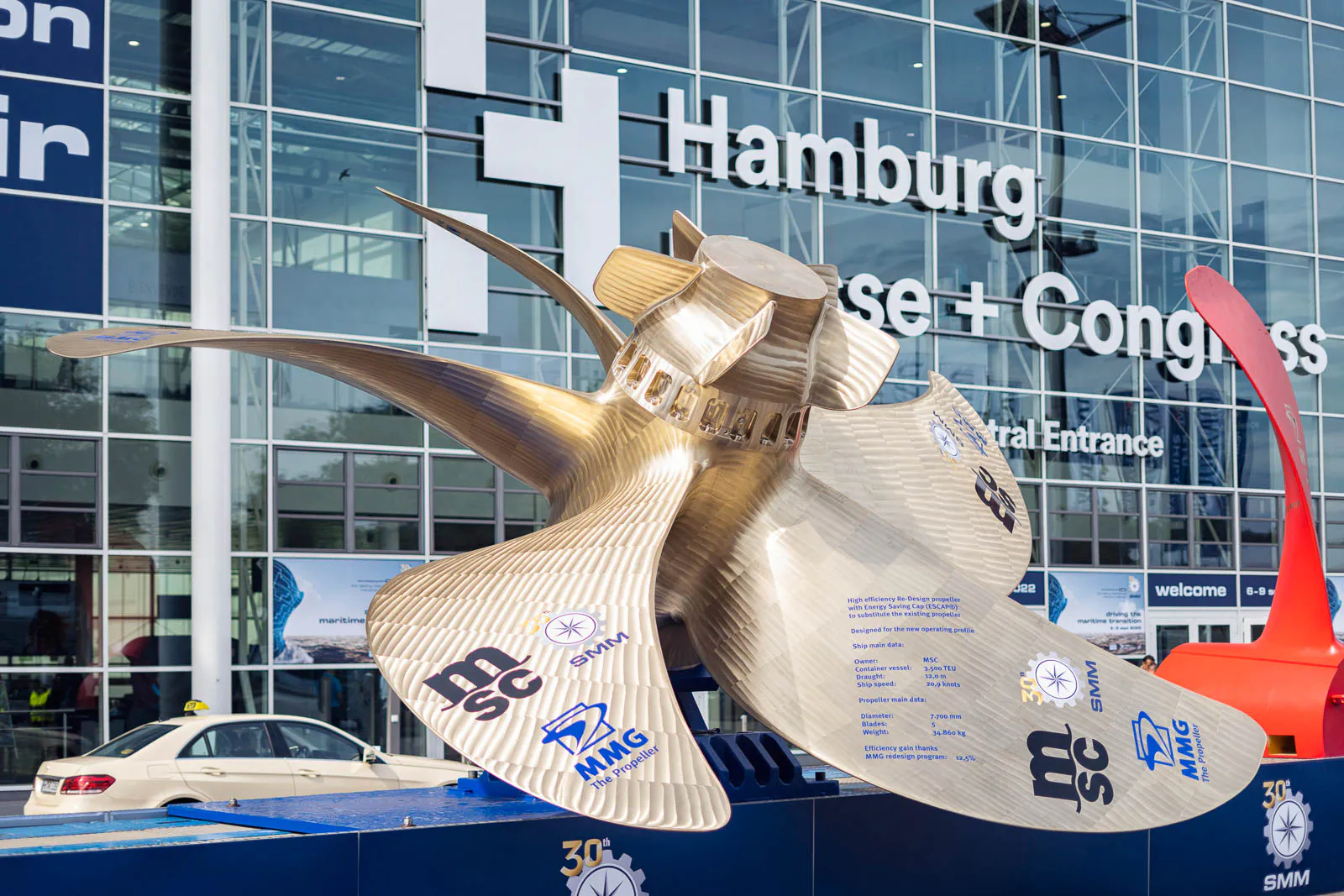 Großer goldfarbener Schiffspropeller vor der Glasfassade der Hamburg Messe, daneben ein roter Propeller.