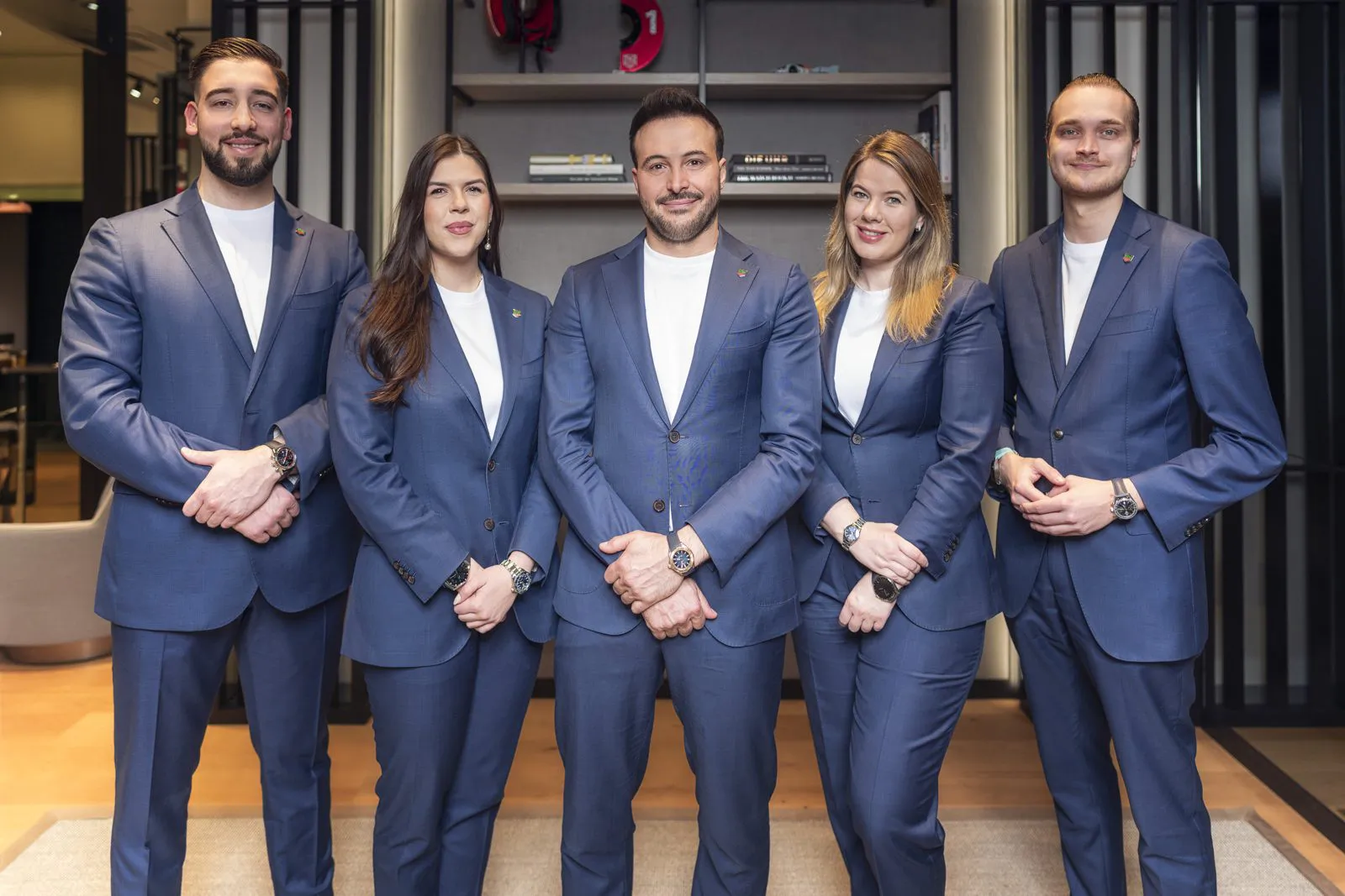 Fünf Tag-Heuer-Mitarbeiter in blauen Anzügen mit Armbanduhren stehen nebeneinander im warm beleuchteten Verkaufsraum.