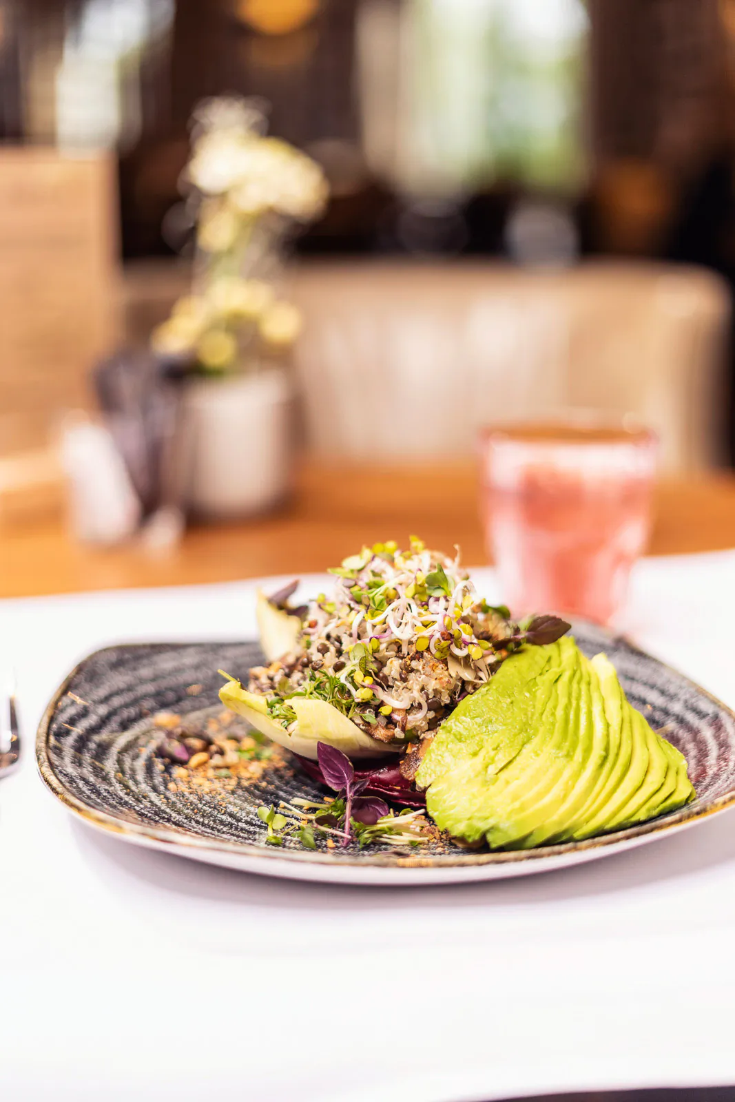 Gefächerte Avocado mit Getreide und Sprossen auf dunklem Teller, dahinter ein rosa Getränk im hellen Restaurant.