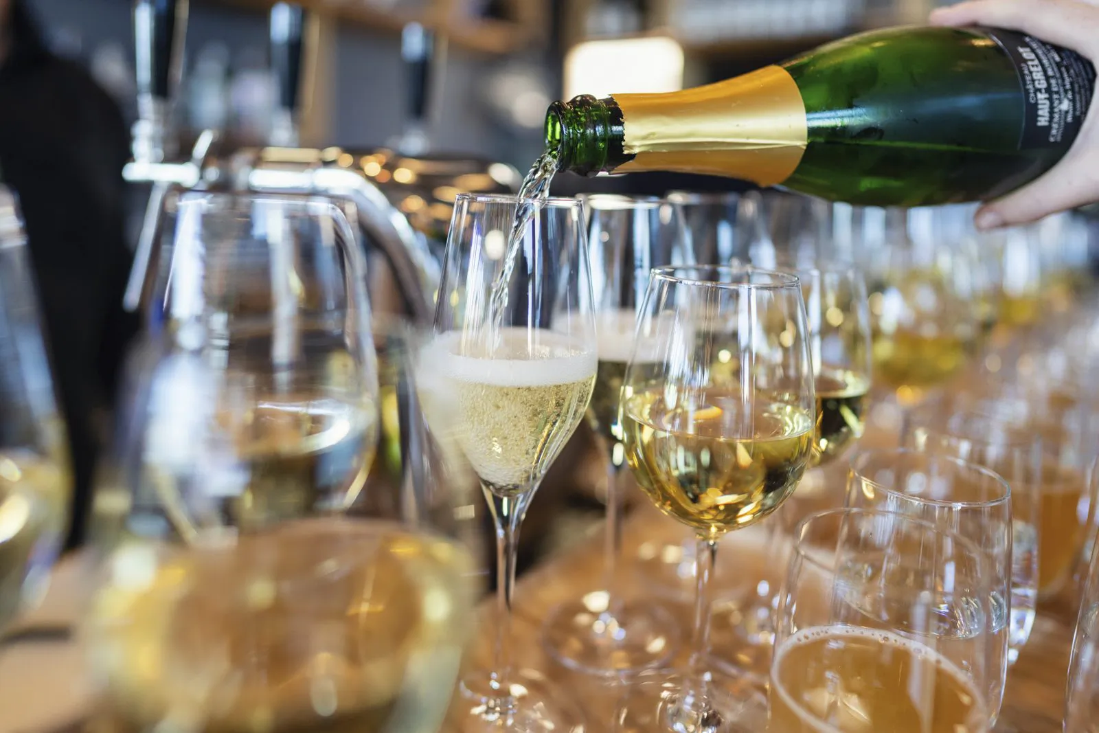 Eine Hand schenkt Sekt aus einer grünen Flasche in ein Glas; auf der Bar stehen zahlreiche gefüllte Gläser.