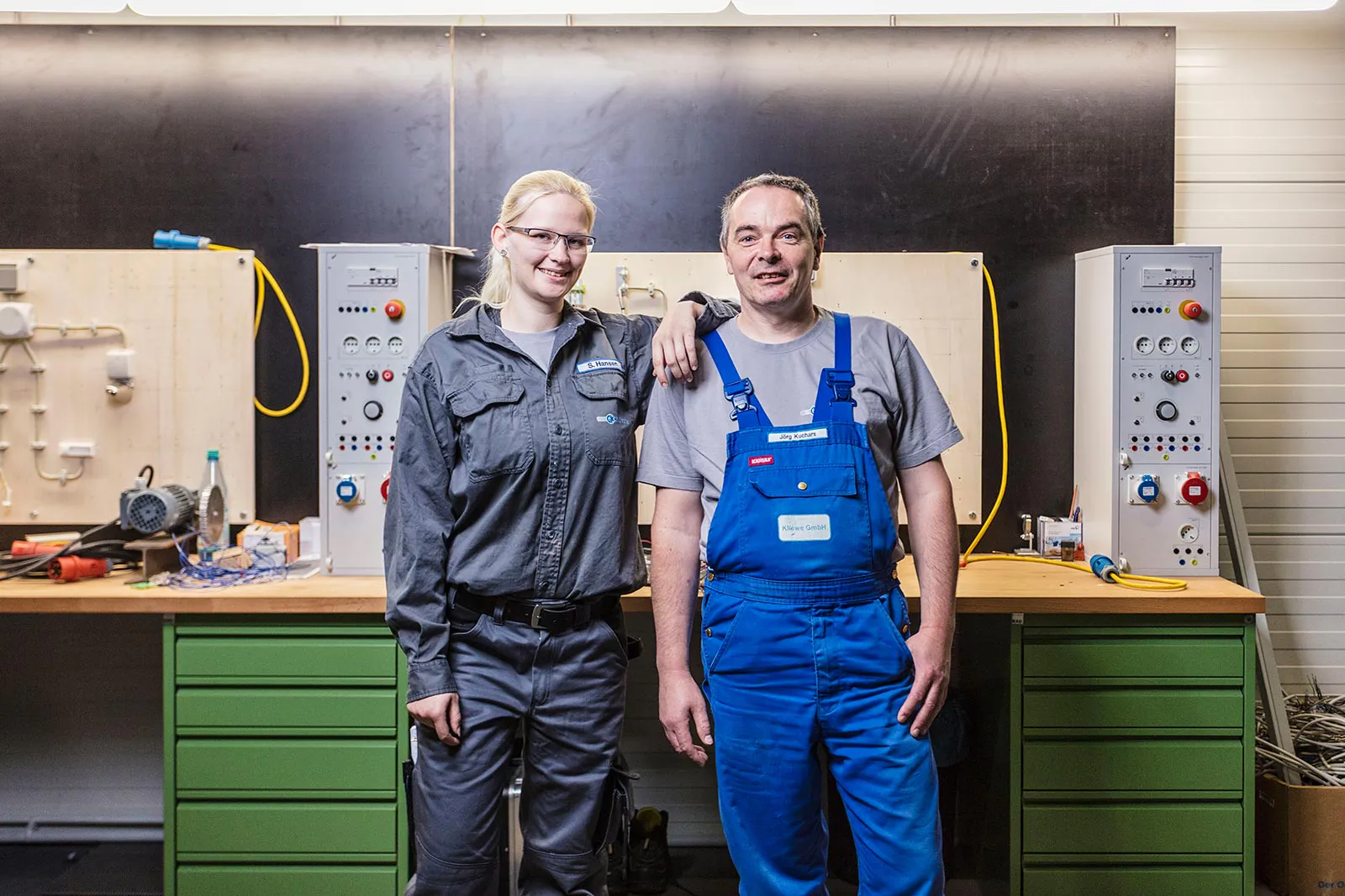 Zwei lächelnde Personen in Arbeitskleidung stehen vor einer Werkbank mit elektrischen Übungsanlagen.