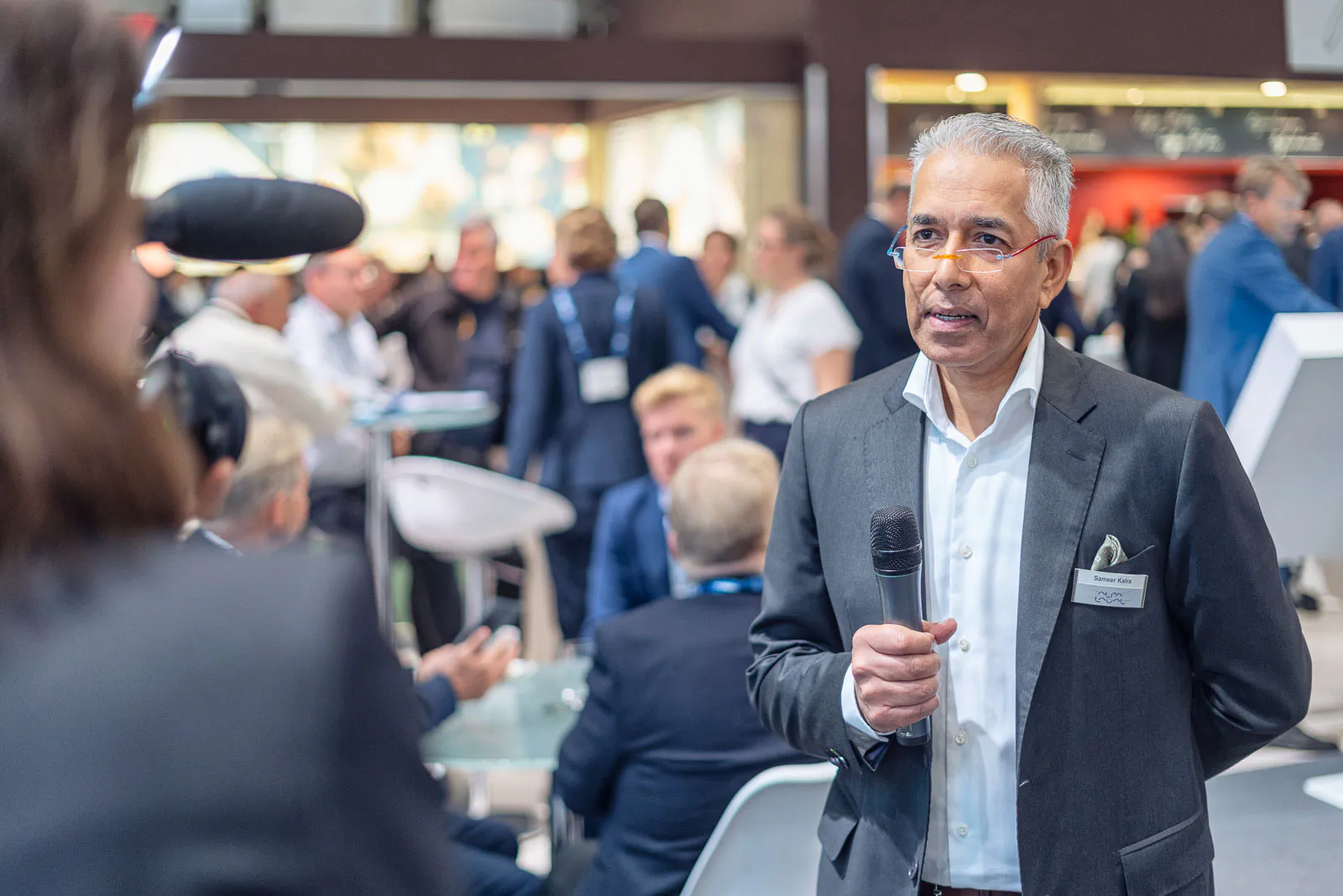 Mann im Anzug mit Mikrofon vor einer Kamera am belebten Messestand von Alfa Laval.