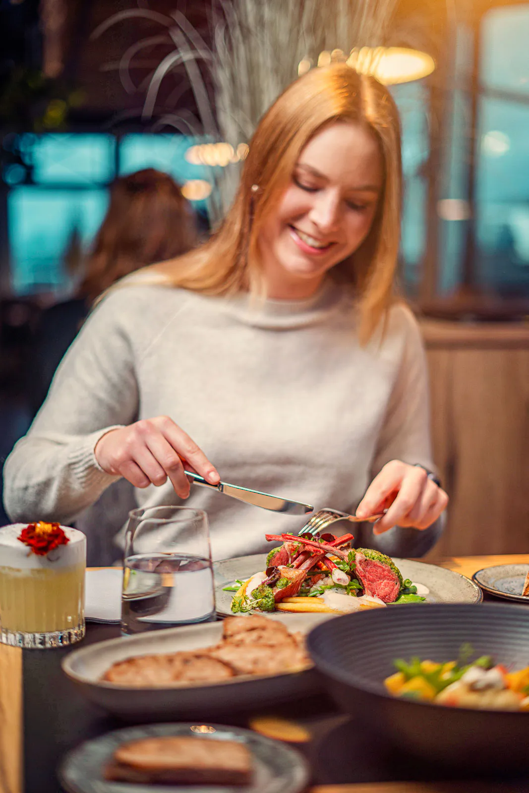 Eine lächelnde Frau schneidet ihr Essen im warm beleuchteten Restaurant, vor ihr stehen Teller und Getränke.
