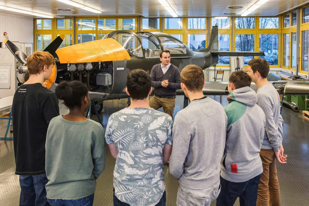 Jugendliche hören einem Mann vor einem Propellerflugzeug mit geöffneter Motorhaube in einer hellen Werkhalle zu.