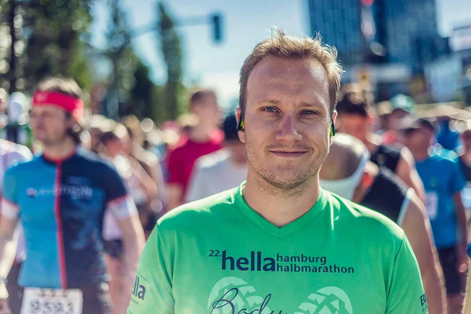 Läufer im grünen Hella-Halbmarathon-Shirt mit Kopfhörern inmitten weiterer Teilnehmer auf sonniger Straße.