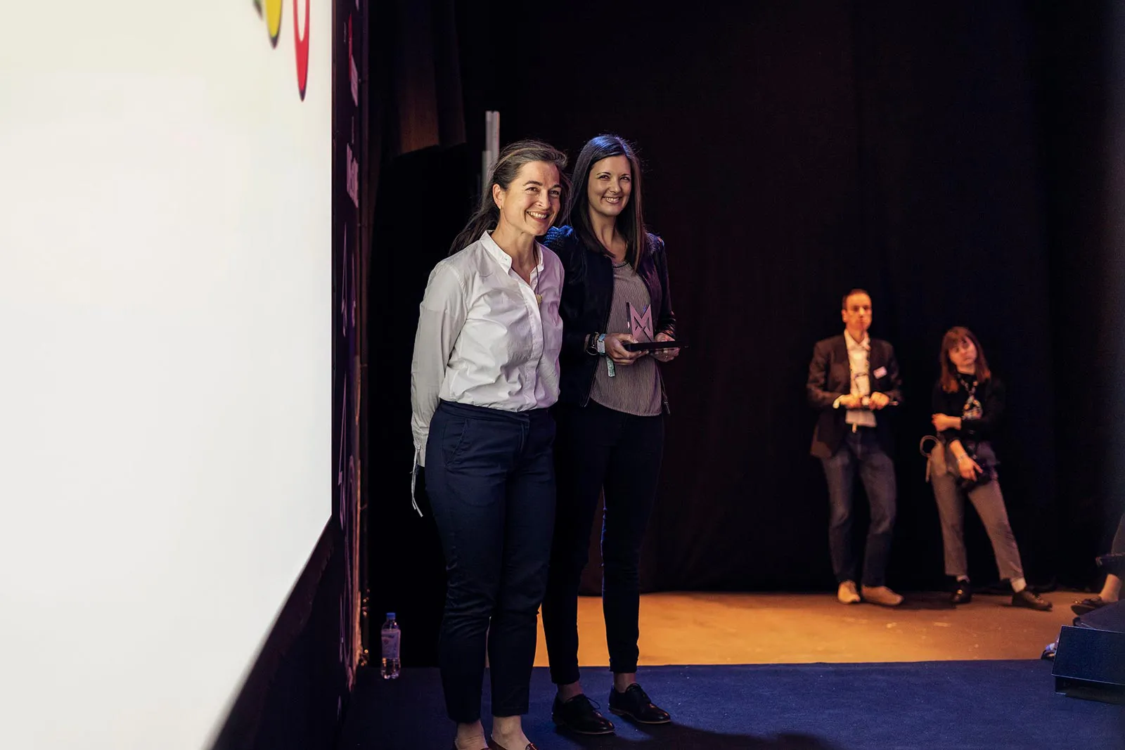 Zwei lächelnde Frauen auf der Bühne beim MAX Award, weitere Personen stehen im Hintergrund.