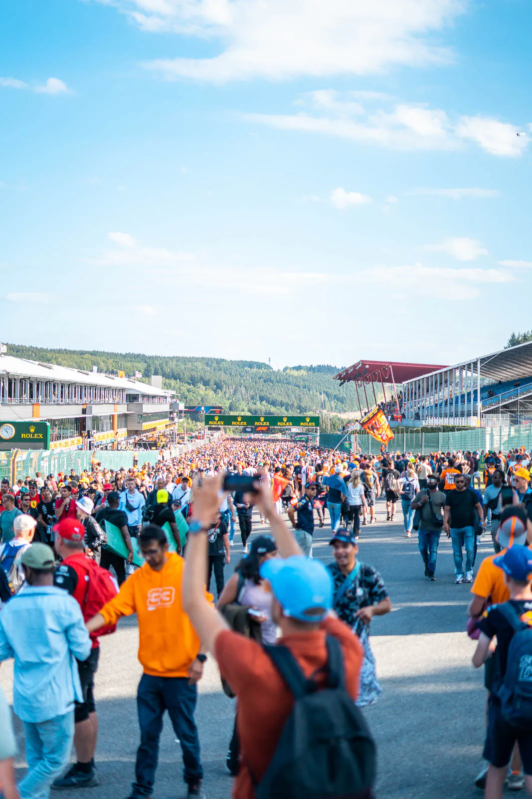 Zahlreiche Fans auf der sonnigen Rennstrecke zwischen Tribünen, im Vordergrund fotografiert ein Mann mit blauer Kappe.