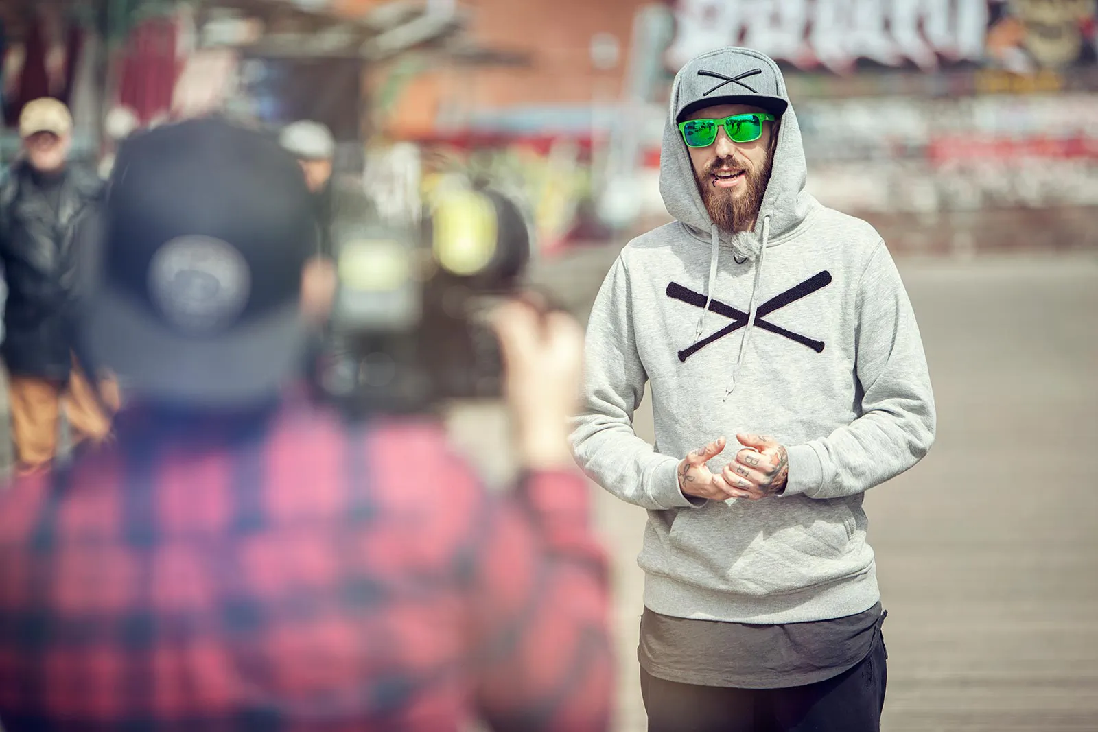 Bärtiger Mann mit grauem Hoodie, Kappe und grüner Sonnenbrille vor einer Videokamera im sonnigen Außenbereich.
