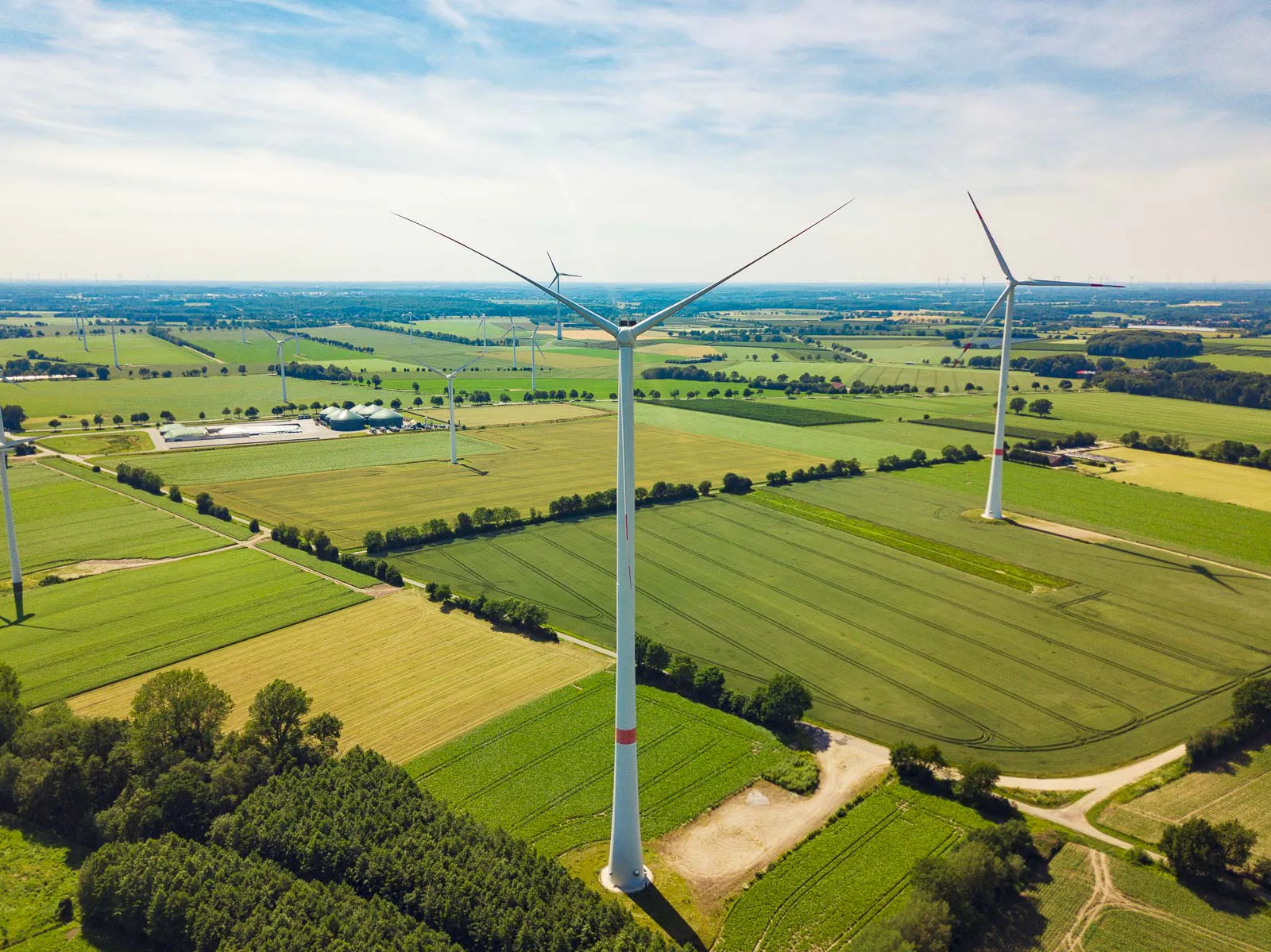 Luftaufnahme mehrerer Windräder zwischen grünen Feldern und Feldwegen bei Tageslicht unter leicht bewölktem Himmel.