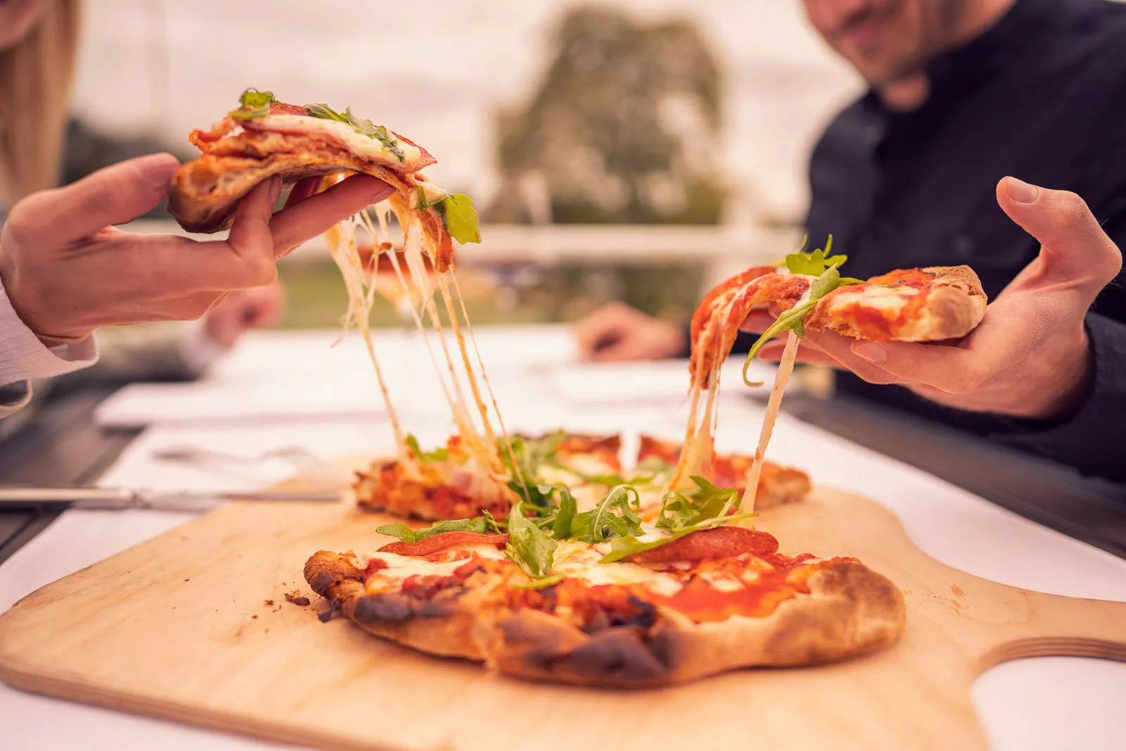 Zwei Personen ziehen Pizzastücke mit langen Käsefäden von einer Pizza mit Rucola auf einem Holzbrett.