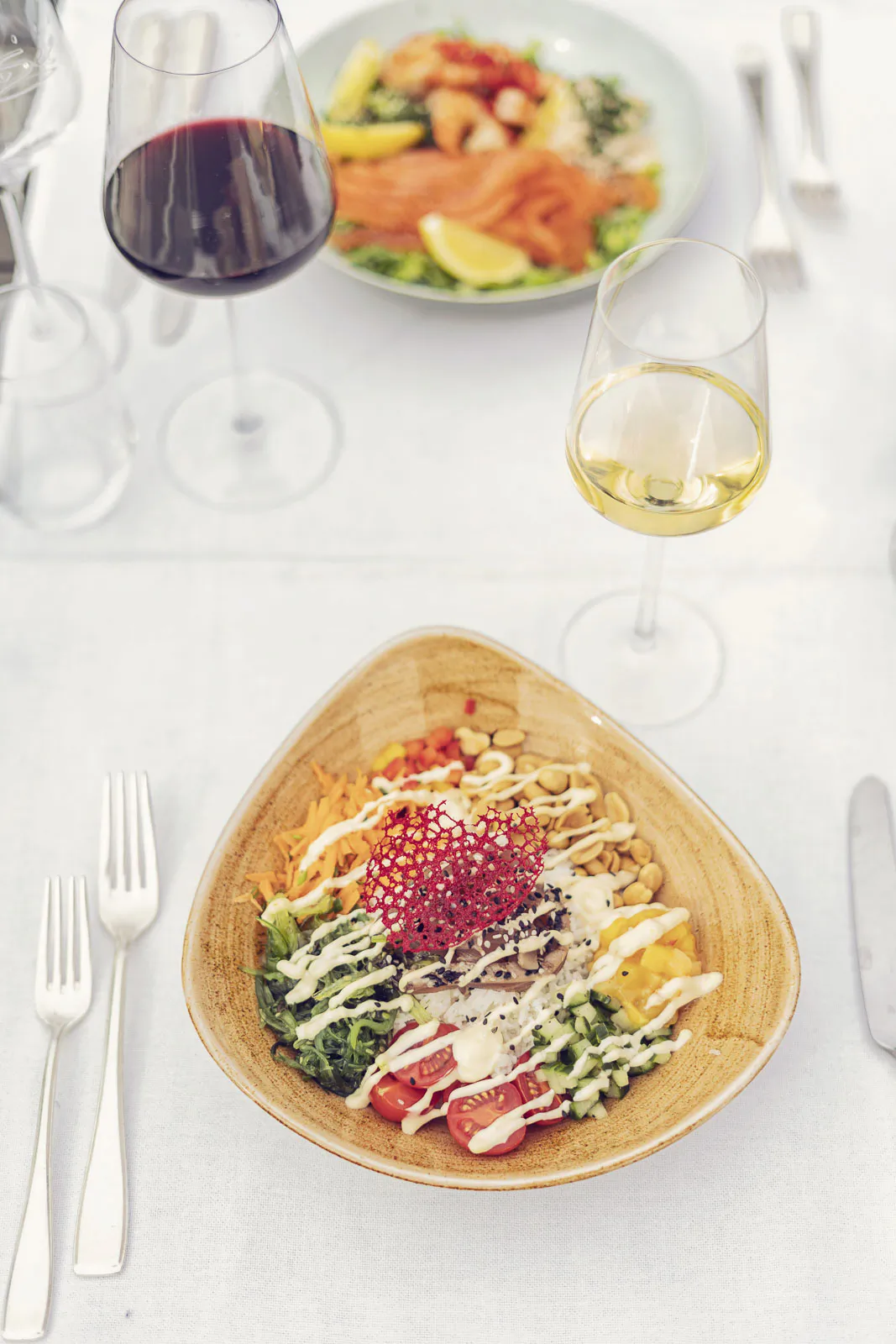 Salatbowl mit Gemüse und Dressing neben Rot- und Weißweingläsern auf einer hell gedeckten Tafel.