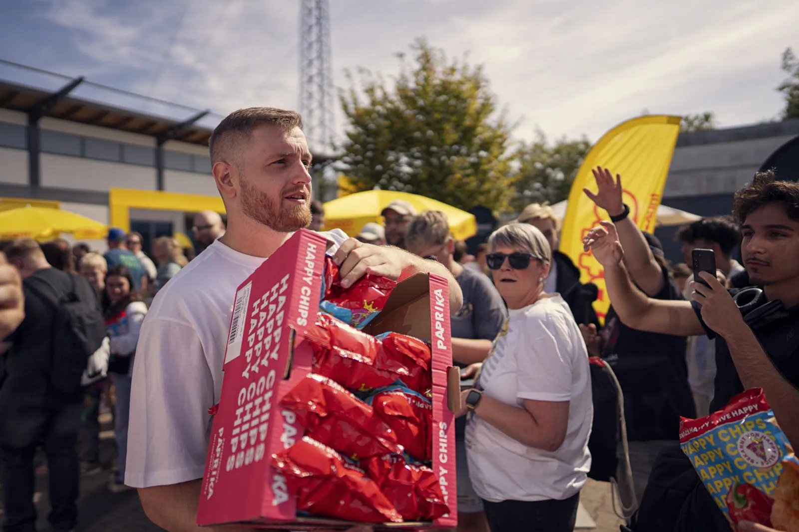 Mann trägt ein rotes Display mit Happy-Chips-Tüten durch eine Menschenmenge bei einem sonnigen Outdoor-Event.
