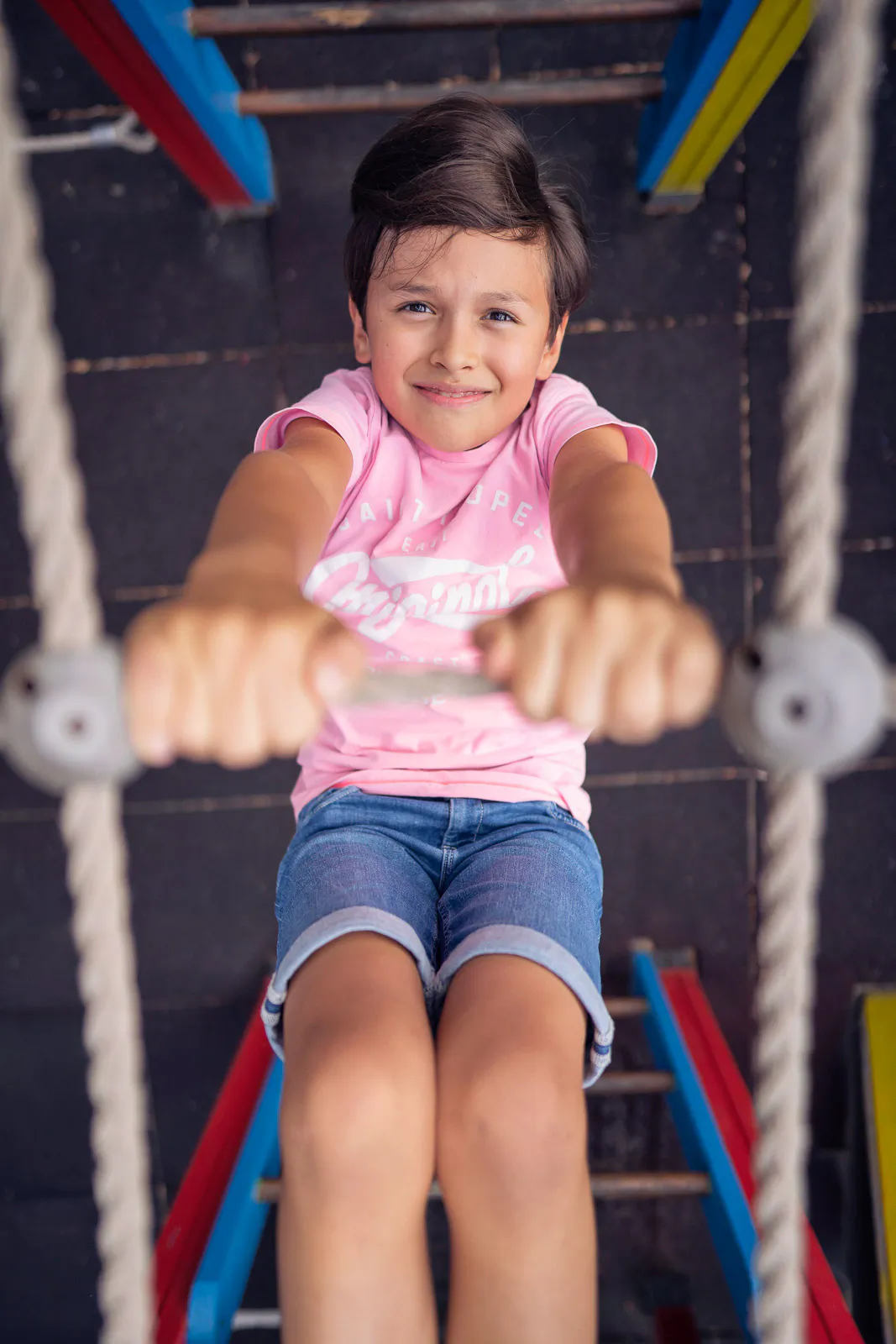 Kind in rosa T-Shirt und Jeansshorts hält sich an einer Turnstange zwischen Seilen fest und blickt in die Kamera.