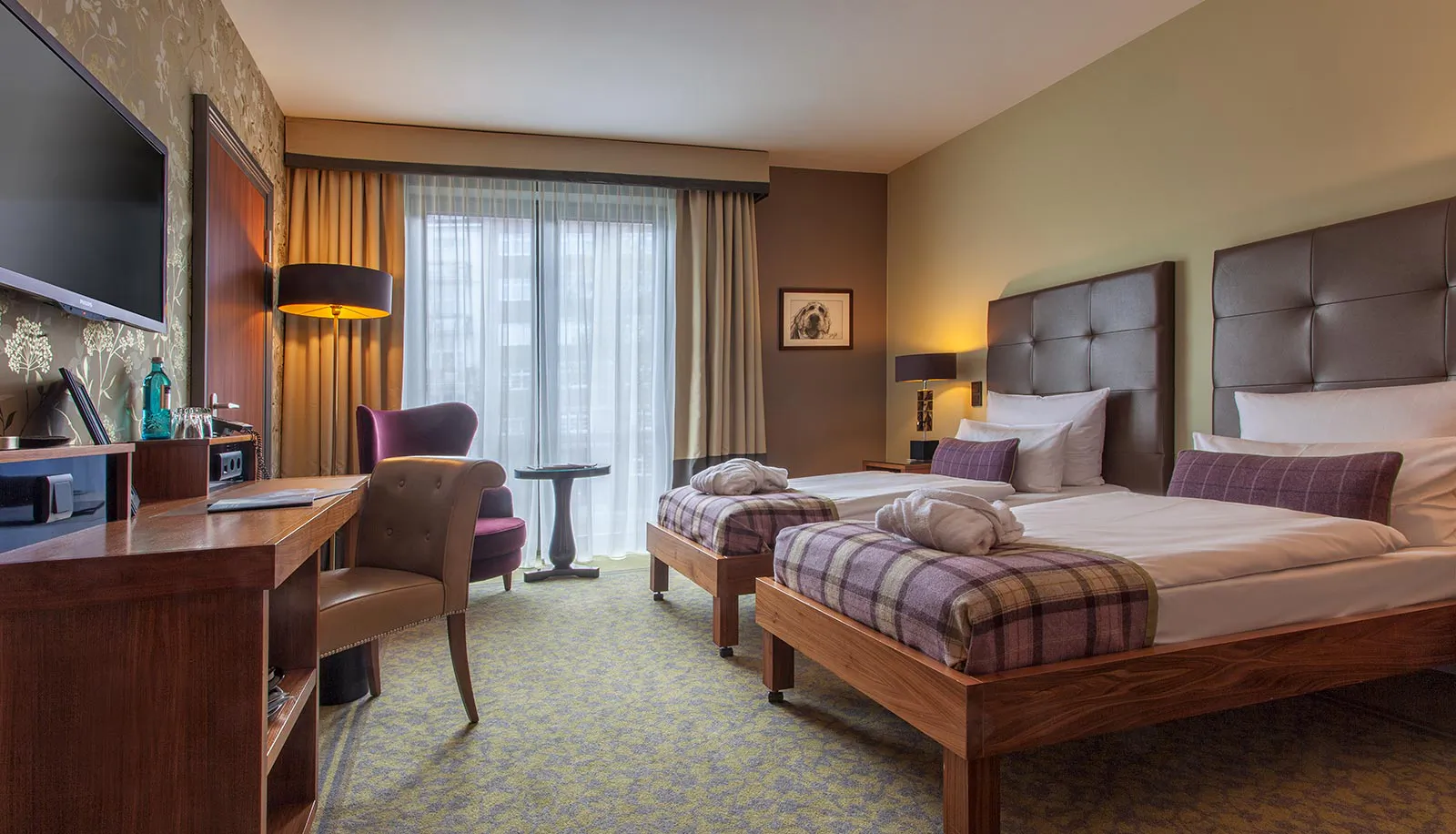 Hotelzimmer mit zwei Einzelbetten, karierten Tagesdecken, Schreibtisch und Sesseln im Tageslicht großer Fenster.