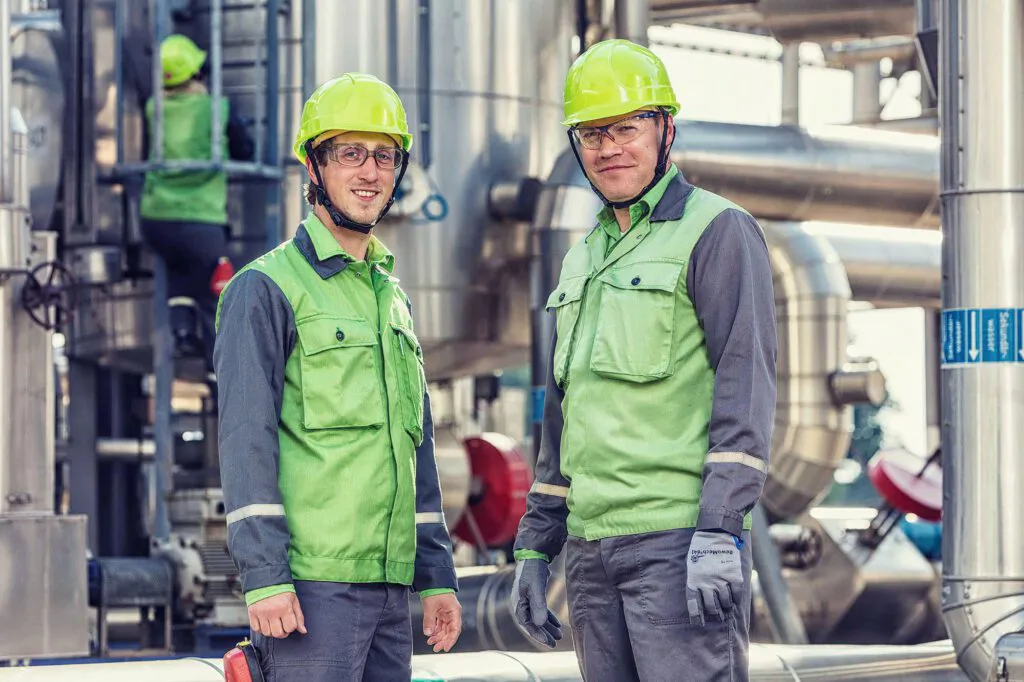 Zwei Mitarbeiter in grüner Schutzkleidung mit Helmen und Schutzbrillen zwischen Rohrleitungen der Raffinerie Heide.