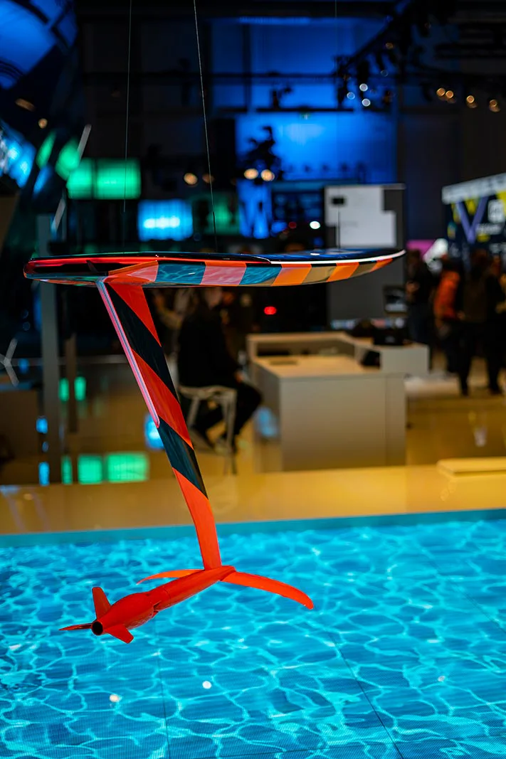 Buntes Hydrofoil-Modell hängt über einer türkis leuchtenden Wasserprojektion in einer Messehalle mit Besuchern.