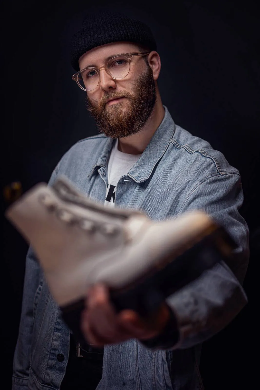 Bärtiger Mann mit Brille, Mütze und Jeansjacke hält einen hellen Dr-Martens-Stiefel vor dunklem Hintergrund.