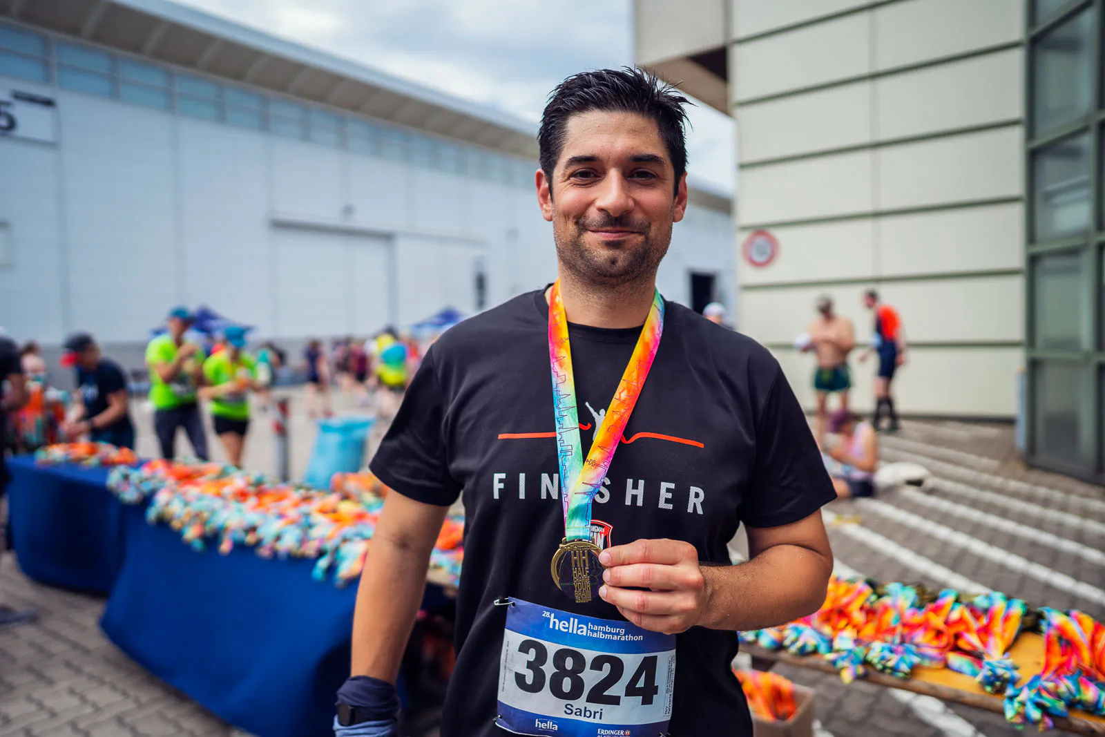 Lächelnder Läufer im schwarzen Finisher-Shirt hält seine Medaille vor Tischen mit bunten Medaillen im Freien.
