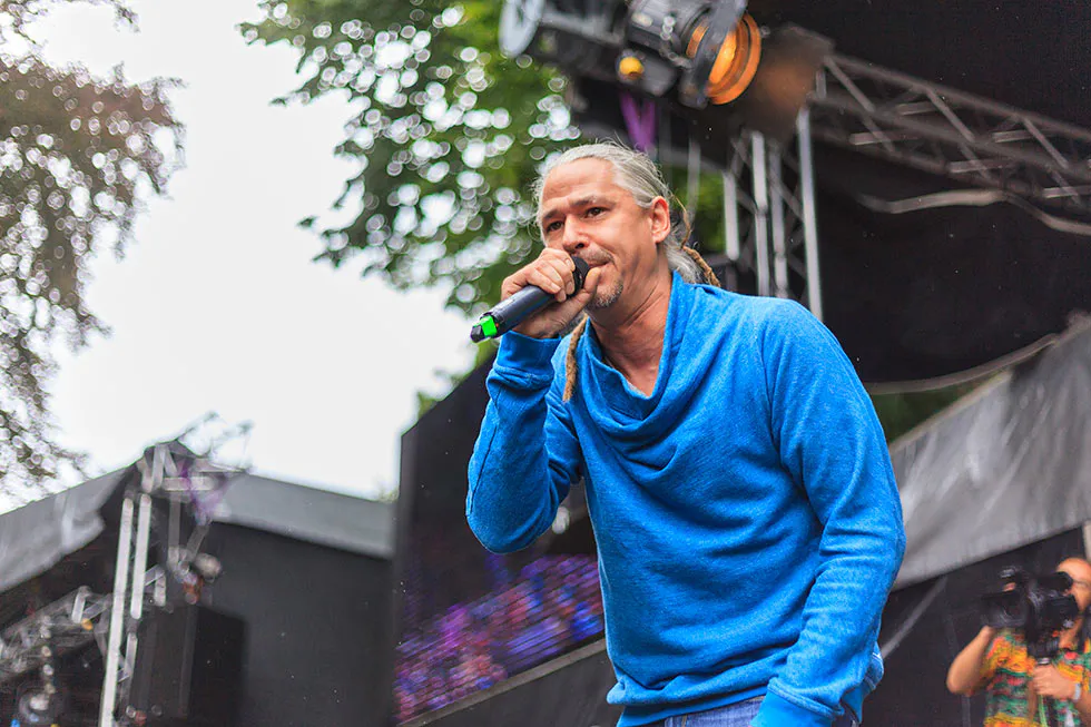 Sänger mit zurückgebundenem Haar und blauem Pullover singt ins Mikrofon auf einer überdachten Festivalbühne.